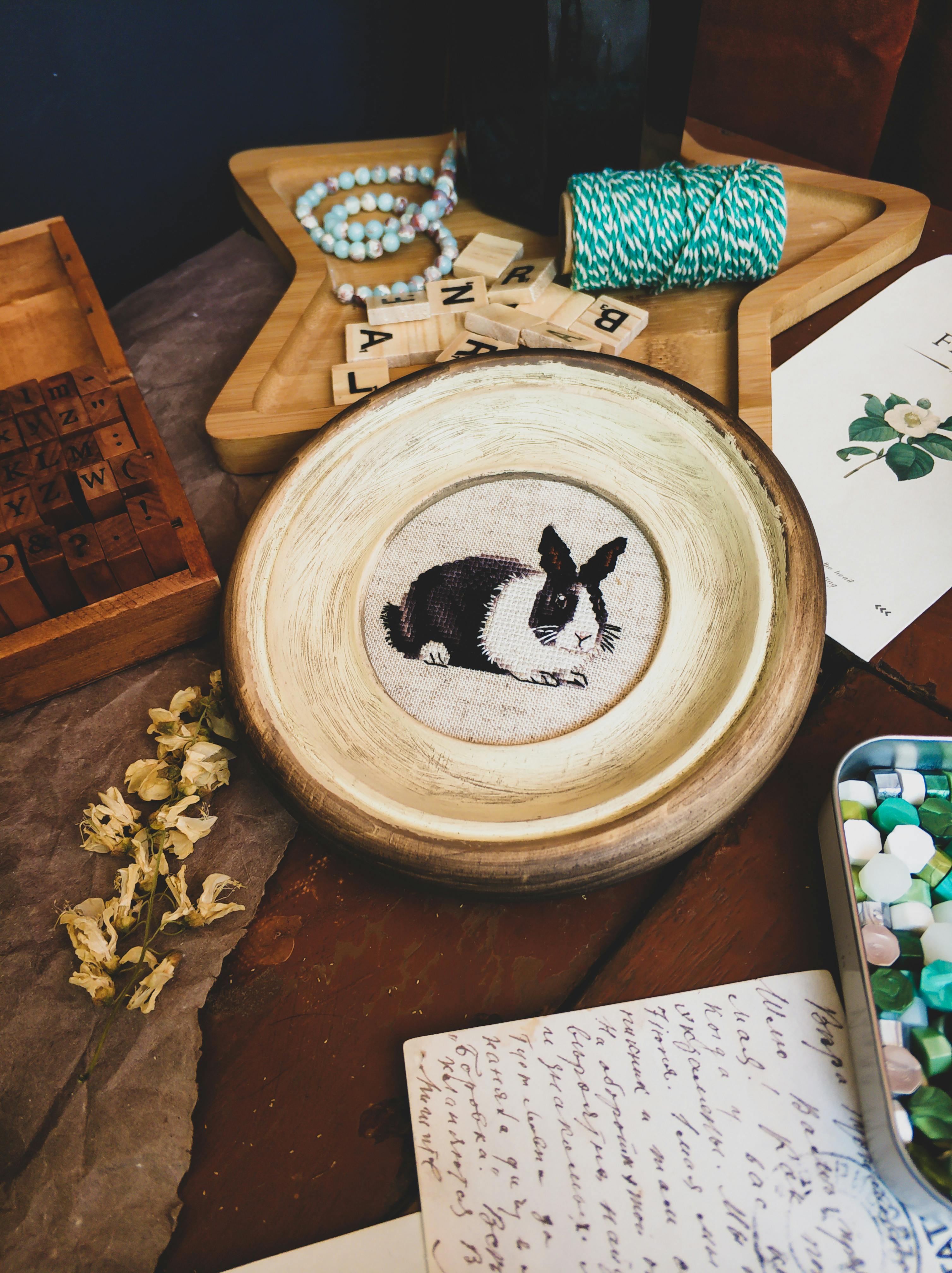 Decorative plate with black and white rabbit painted in the center of it with wooden and metal containers filled with beads and letter blocks placed on a wooden table
