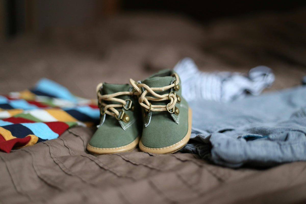 Green and beige baby sneakers with an outfit laying next to them on a brown textile