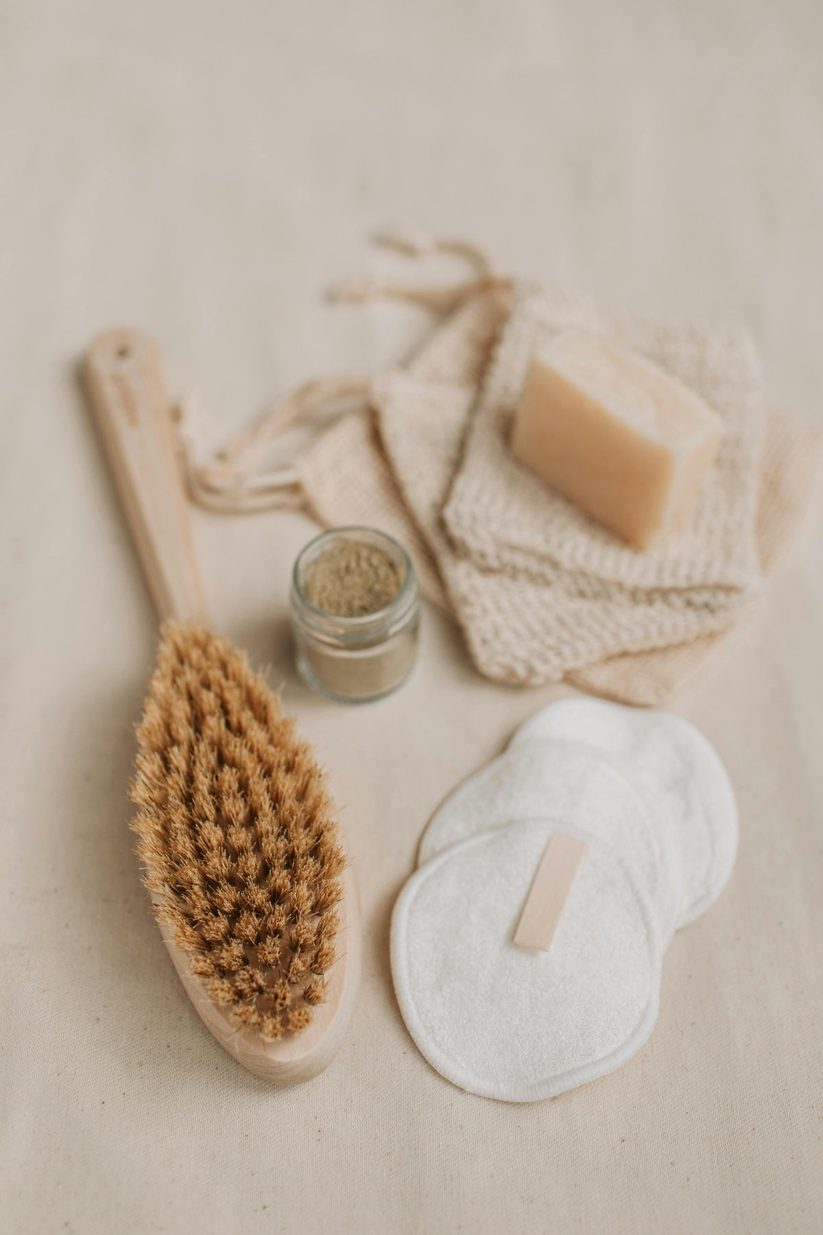 Eco-friendly personal hygiene products, including a wooden body brush for drying, reusable cotton pads, a bar of soap and a small jar of scrub.