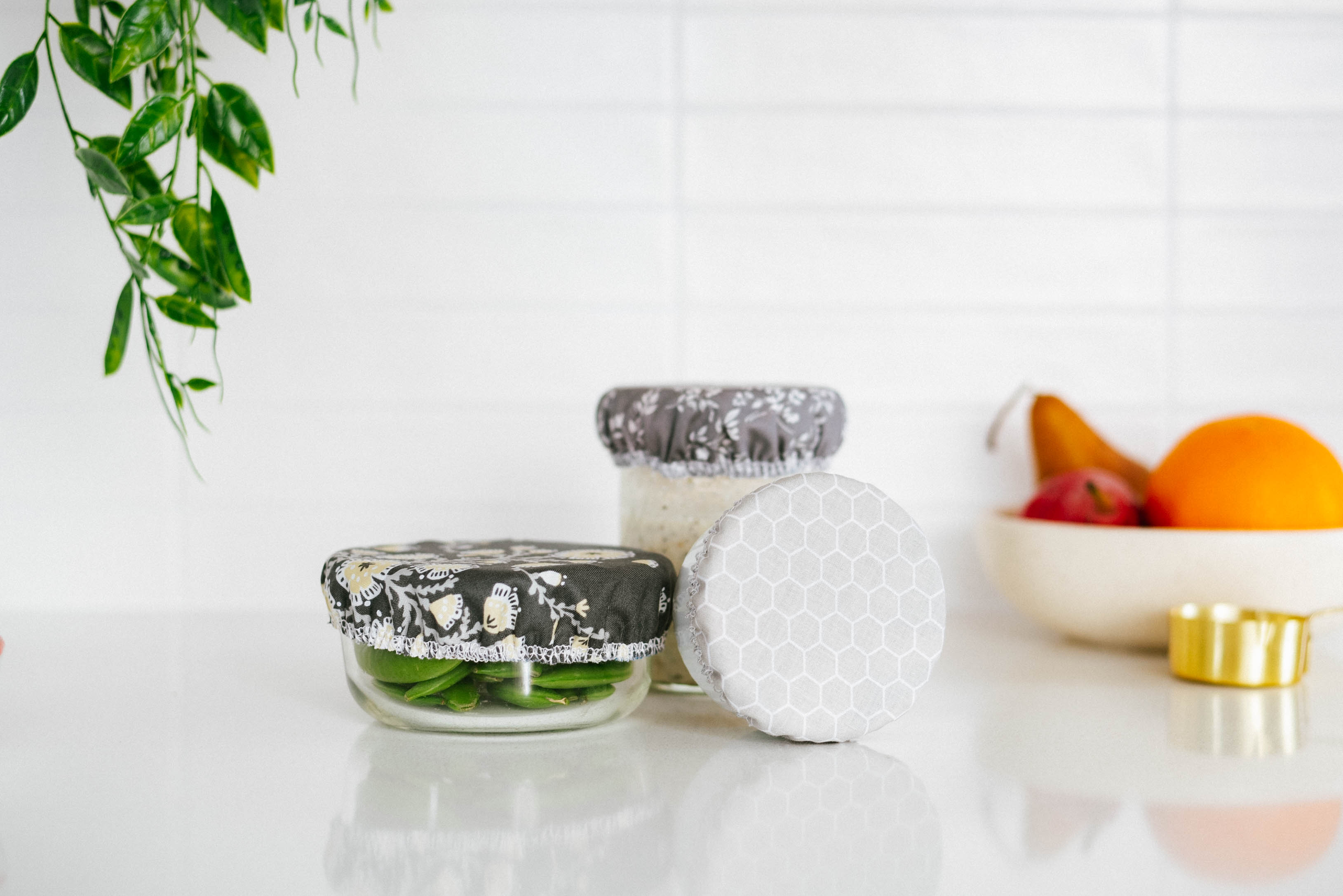 Reusable containers with fabric lids on a white surface with a plant and bowl of fruit in the background.