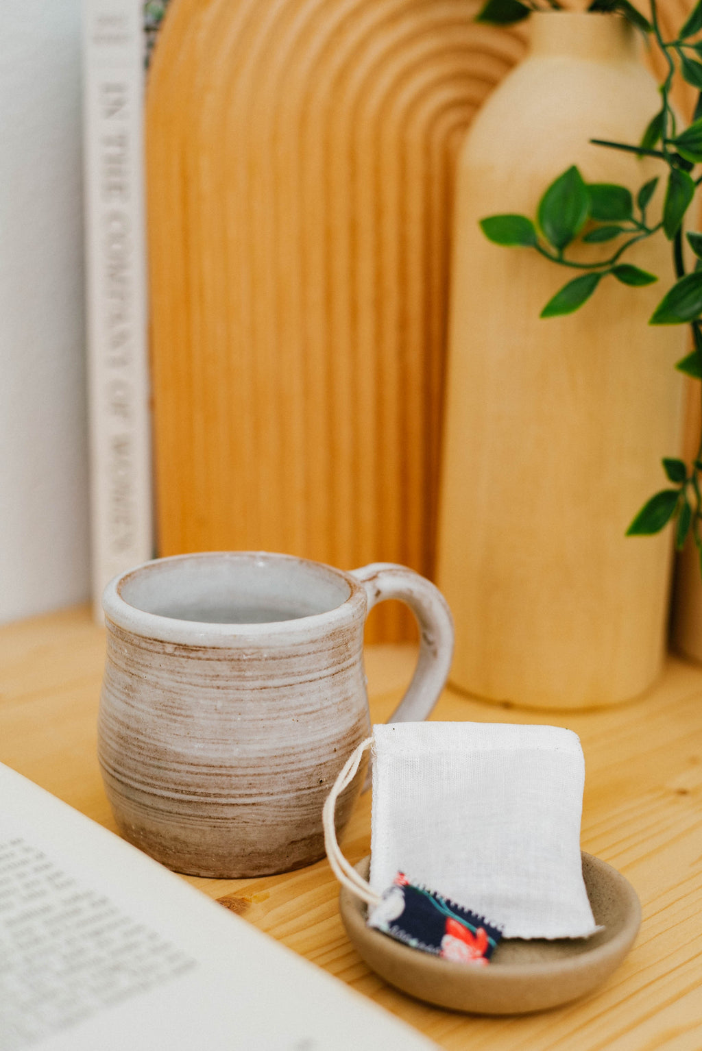 Ceramic mug and small dish with a tea bag on a wooden surface, with a blurred vase and plant in the background.