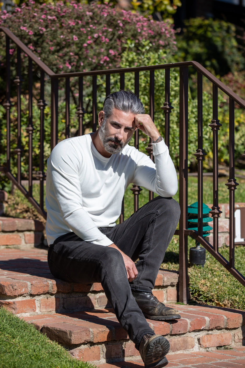 Man wearing long sleeve white t-shirt on steps outdoors with a garden in the background
