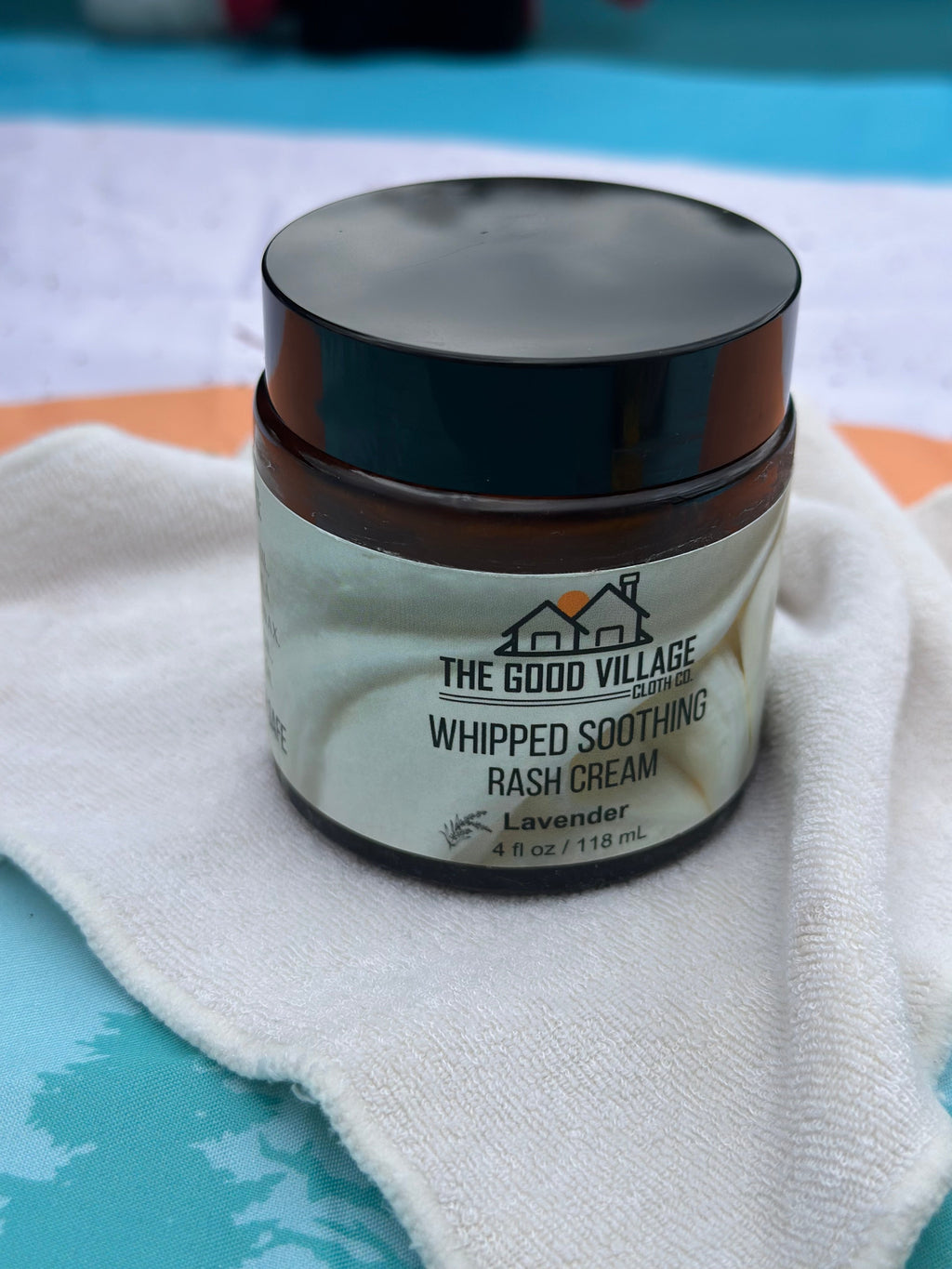 Jar of 'Whipped Soothing Rash Cream' from 'The Good Village' on a white cloth with a blue background.