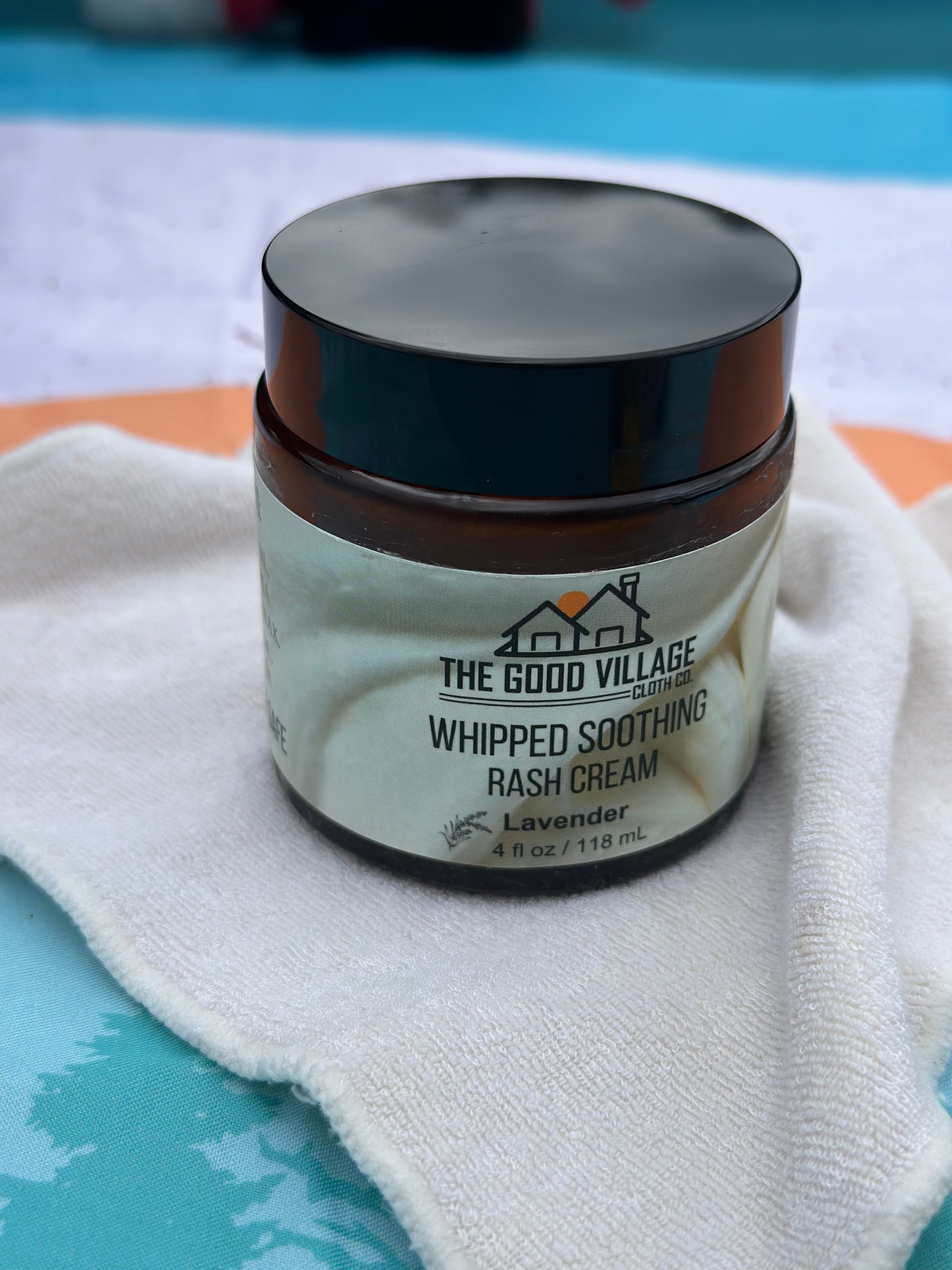 Jar of 'Whipped Soothing Rash Cream' from 'The Good Village' on a white cloth with a blue background.