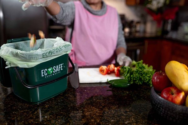 Person using an Ecosafe food waste bin in a kitchen setting with fruits and vegetables.