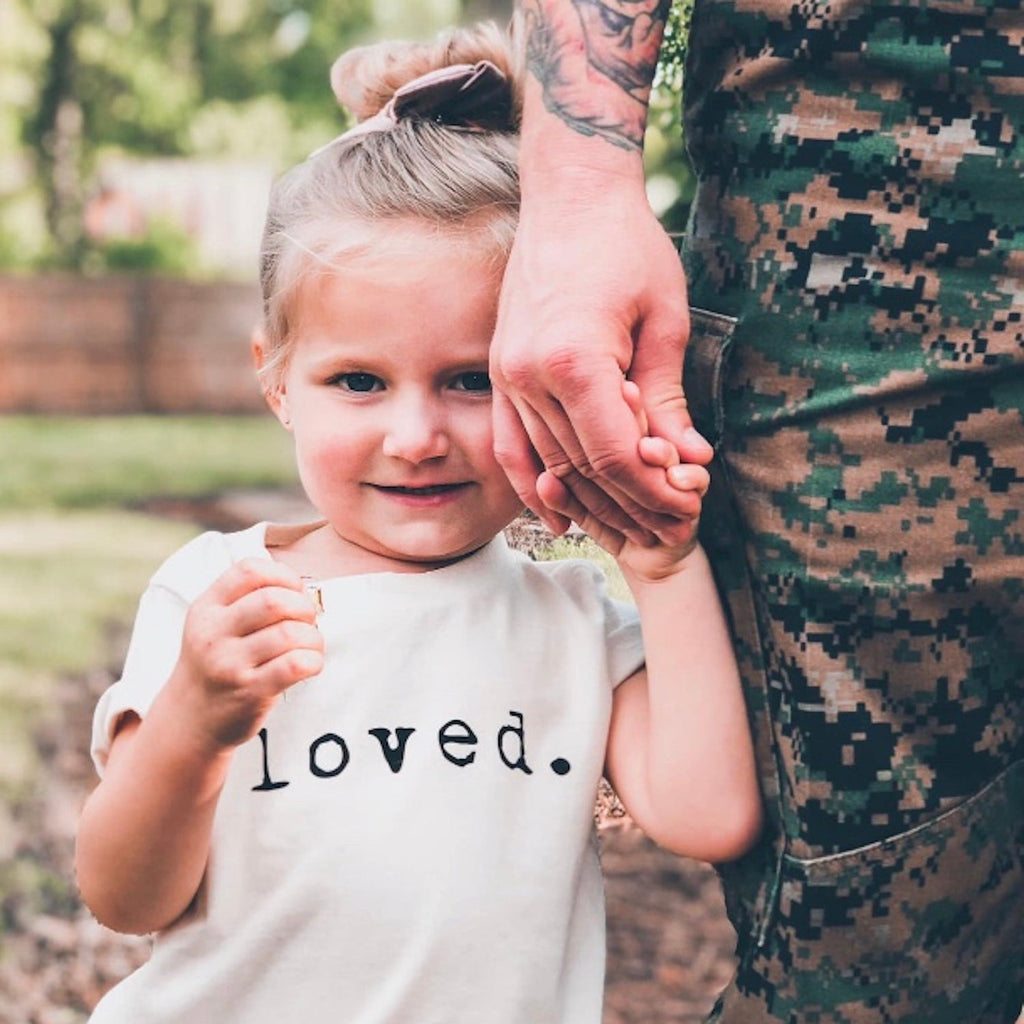 Child wearing a shirt that says 'loved' holding hands with a person in camouflage clothing outdoors.