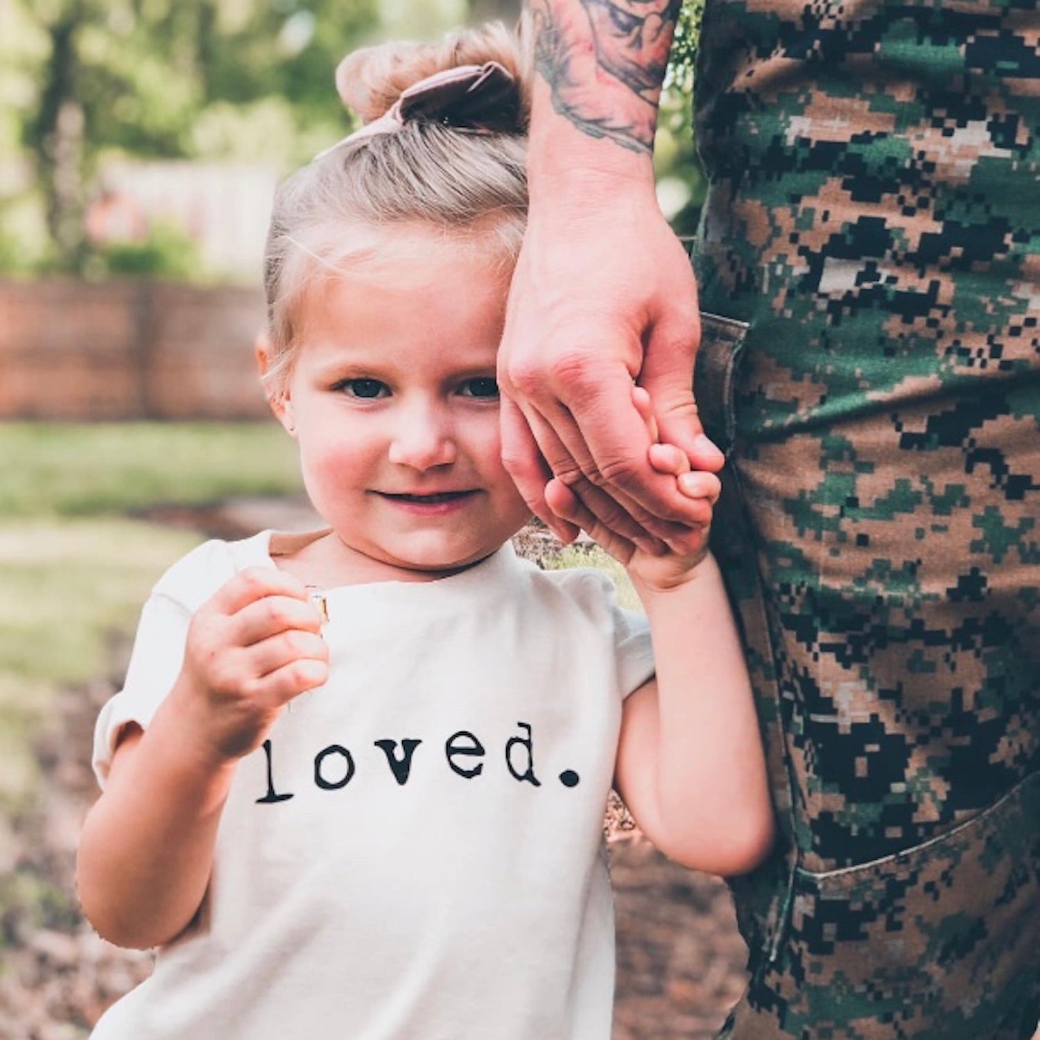 Child wearing a shirt that says 'loved' holding hands with a person in camouflage clothing outdoors.