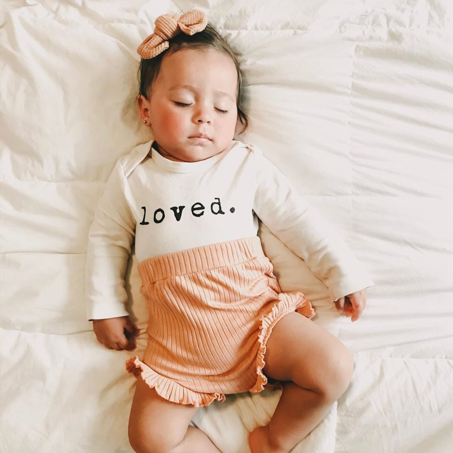 Baby wearing a 'loved' shirt and pink shorts lying on a white bed.