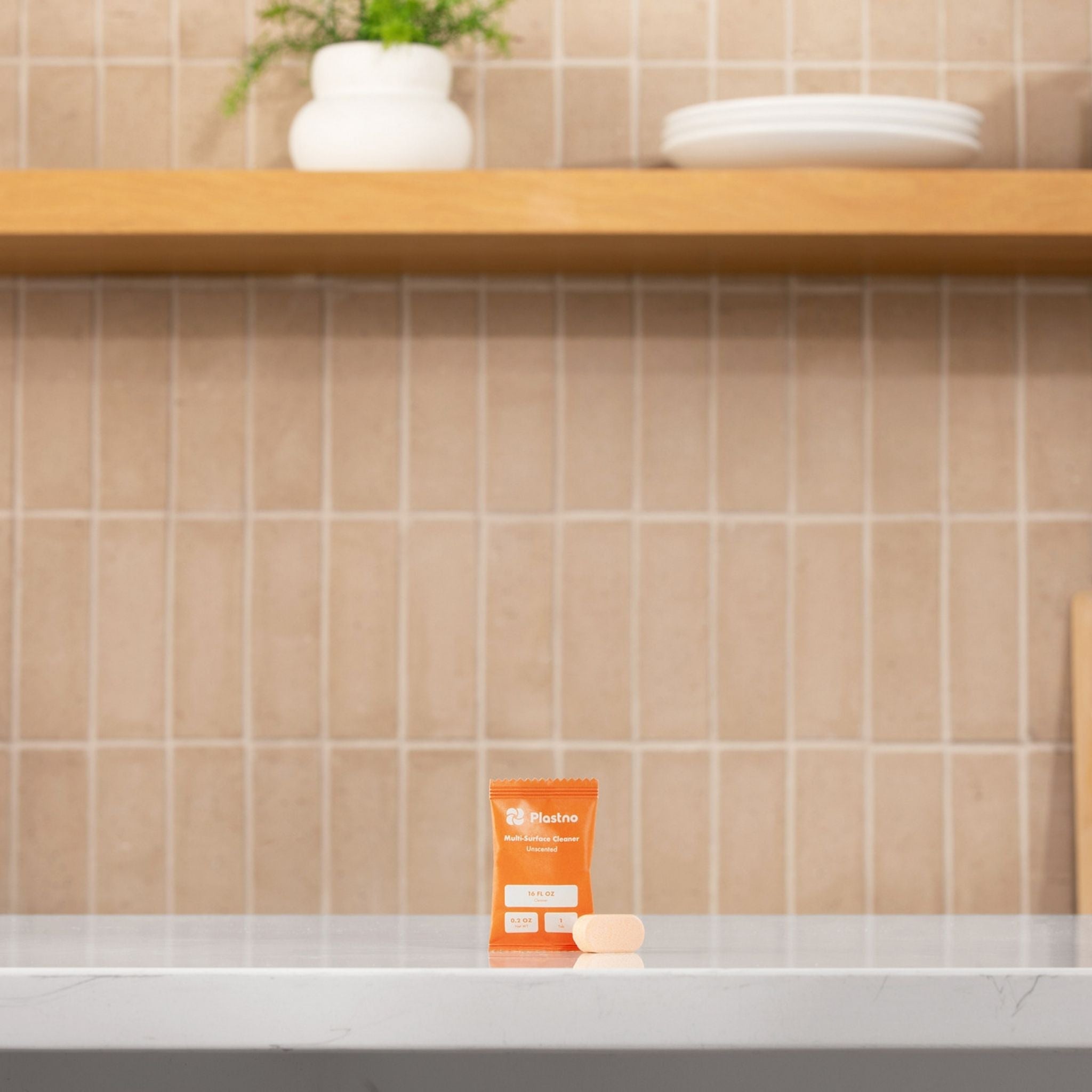 Orange multi-cleaner packet on a kitchen counter with tiled wall and wooden shelf in the background