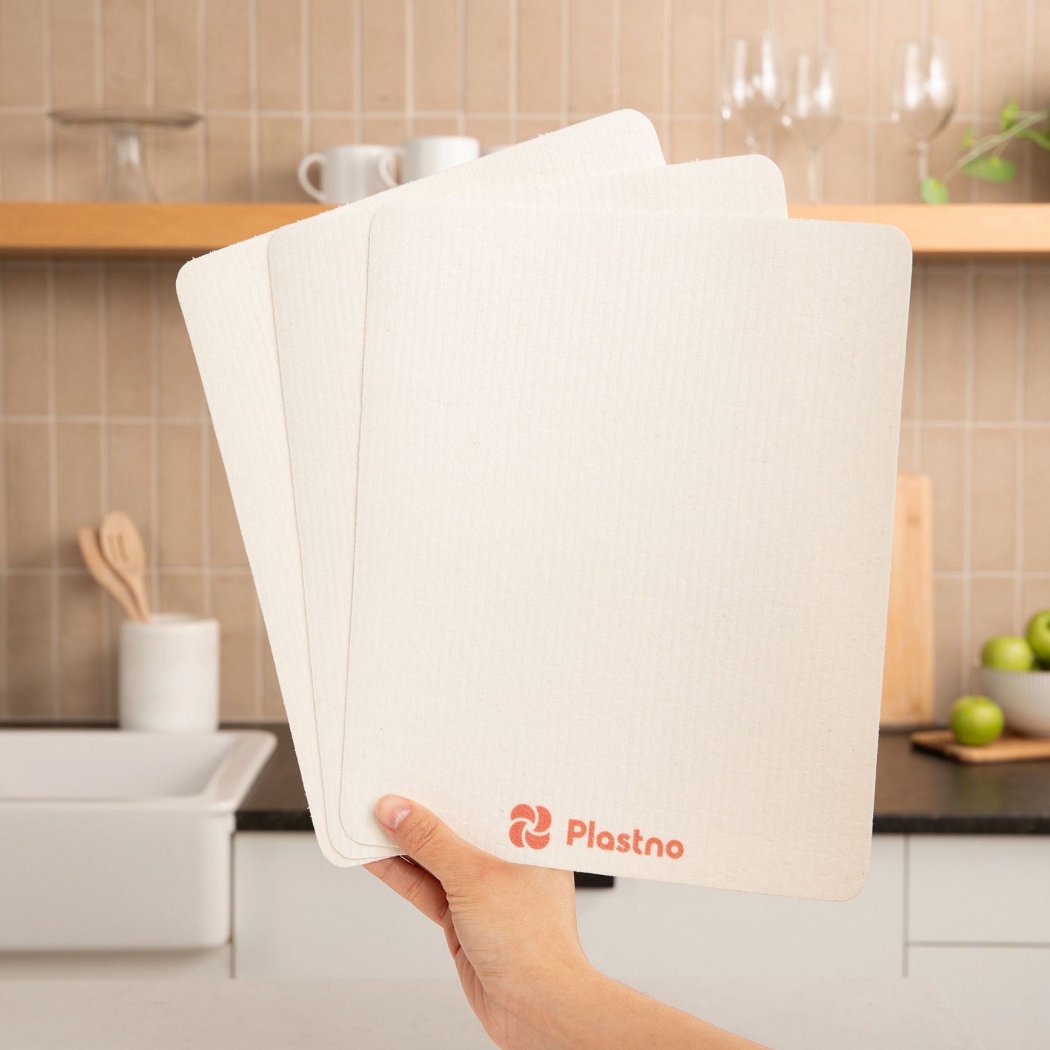 Hand holding three white kitchen towels with Plastino logo in a kitchen setting