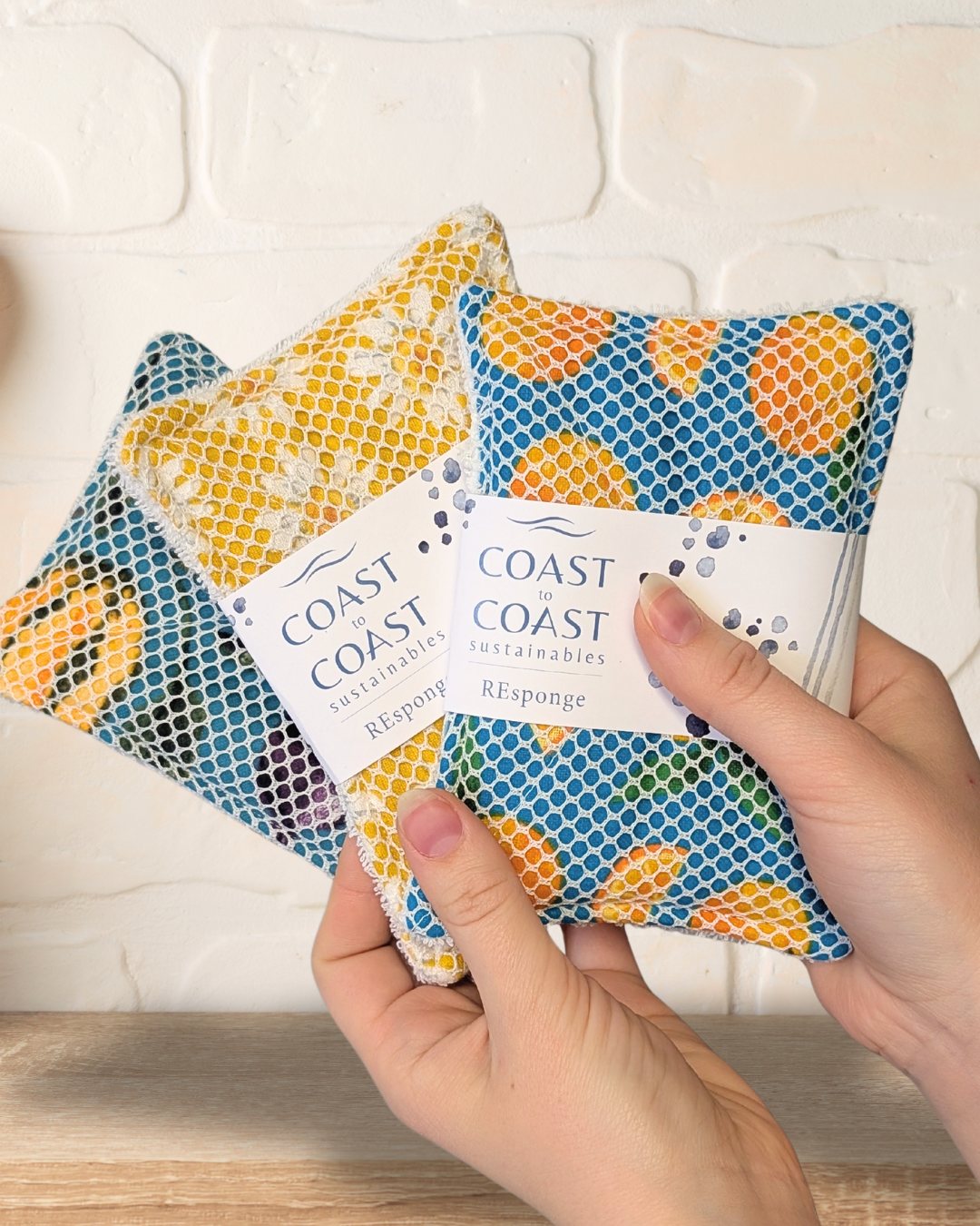 Hand holding three reusable cleaning sponges with 'Coast Coast' branding against a white tiled wall.