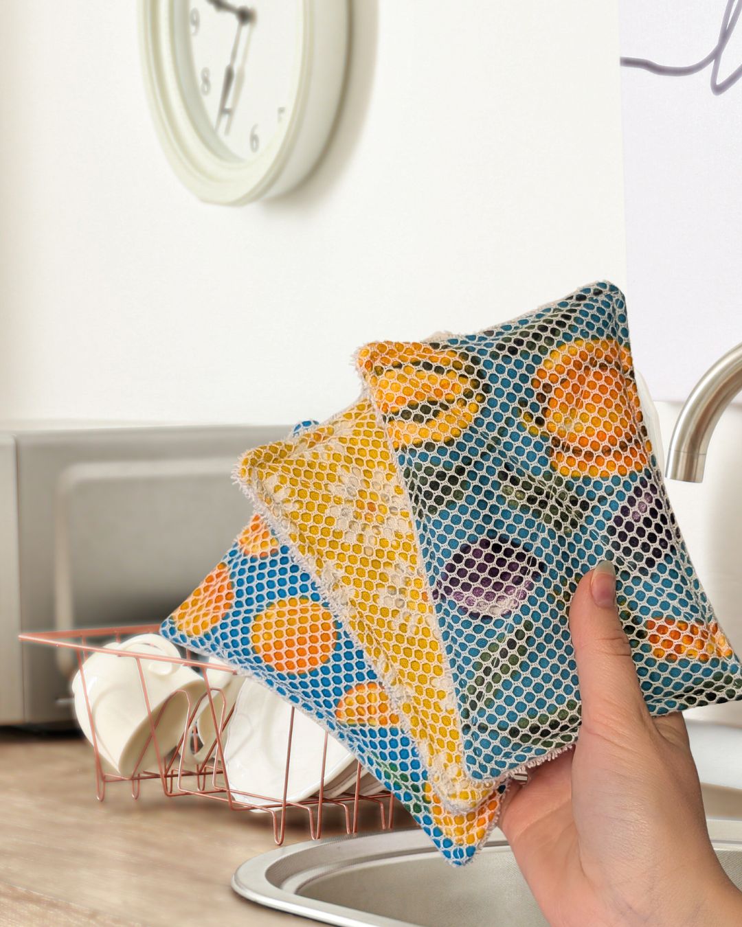 Hand holding three colorful patterned reusable dish sponges in a kitchen setting with a clock and dish rack in the background.