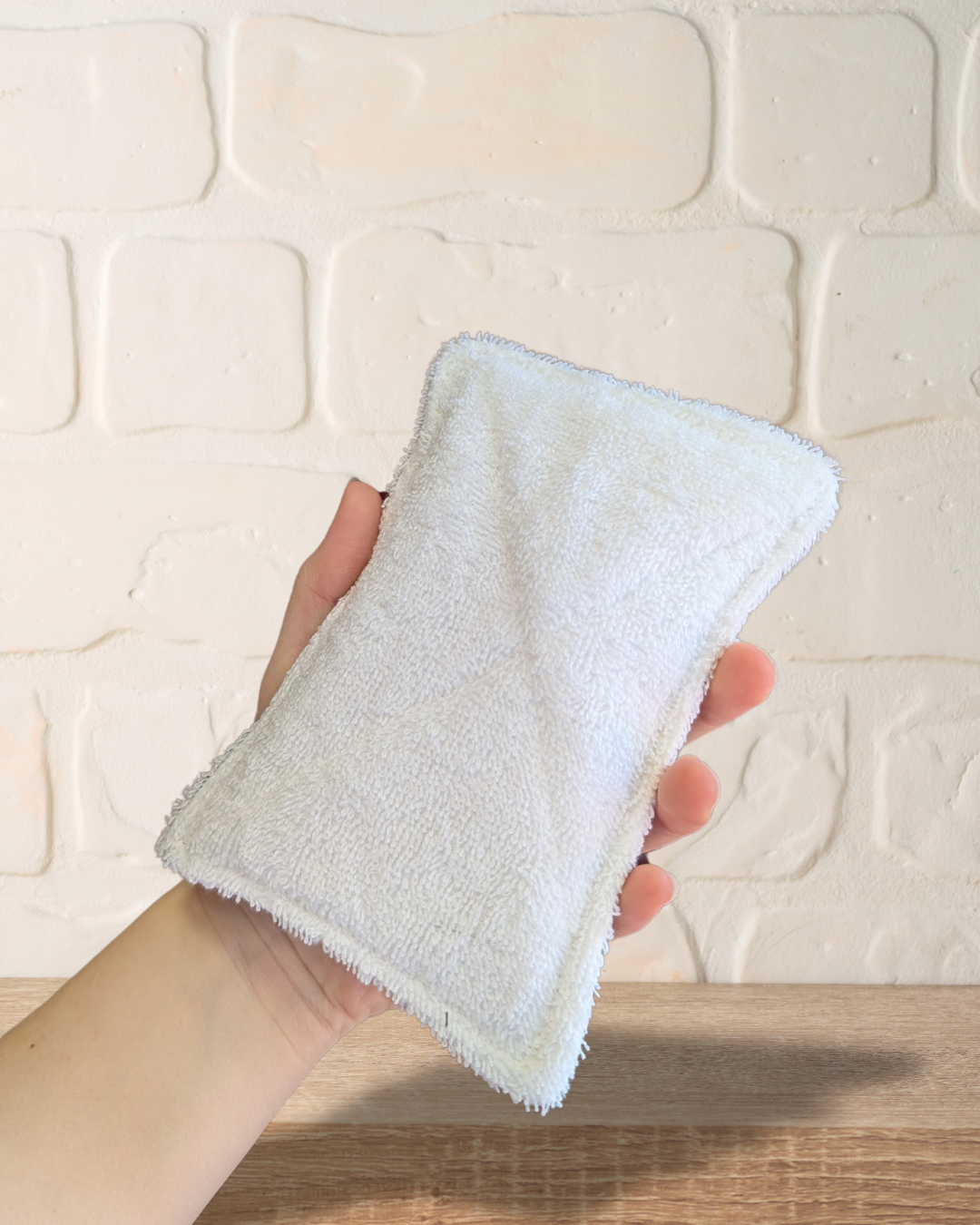 Hand holding a white reusable dish sponge against a textured wall background