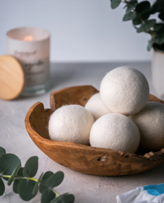 White spherical laundry balls in a wooden bowl with a candle and plant in the background.