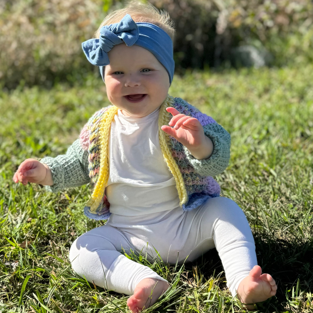 Toddler sitting on grass outdoors wearing ivory bamboo romper and knitted sweater vest
