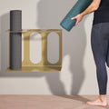 Person holding a yoga mat next to a gold yoga mat stand, attached to the wall with 3 places to hold mats.