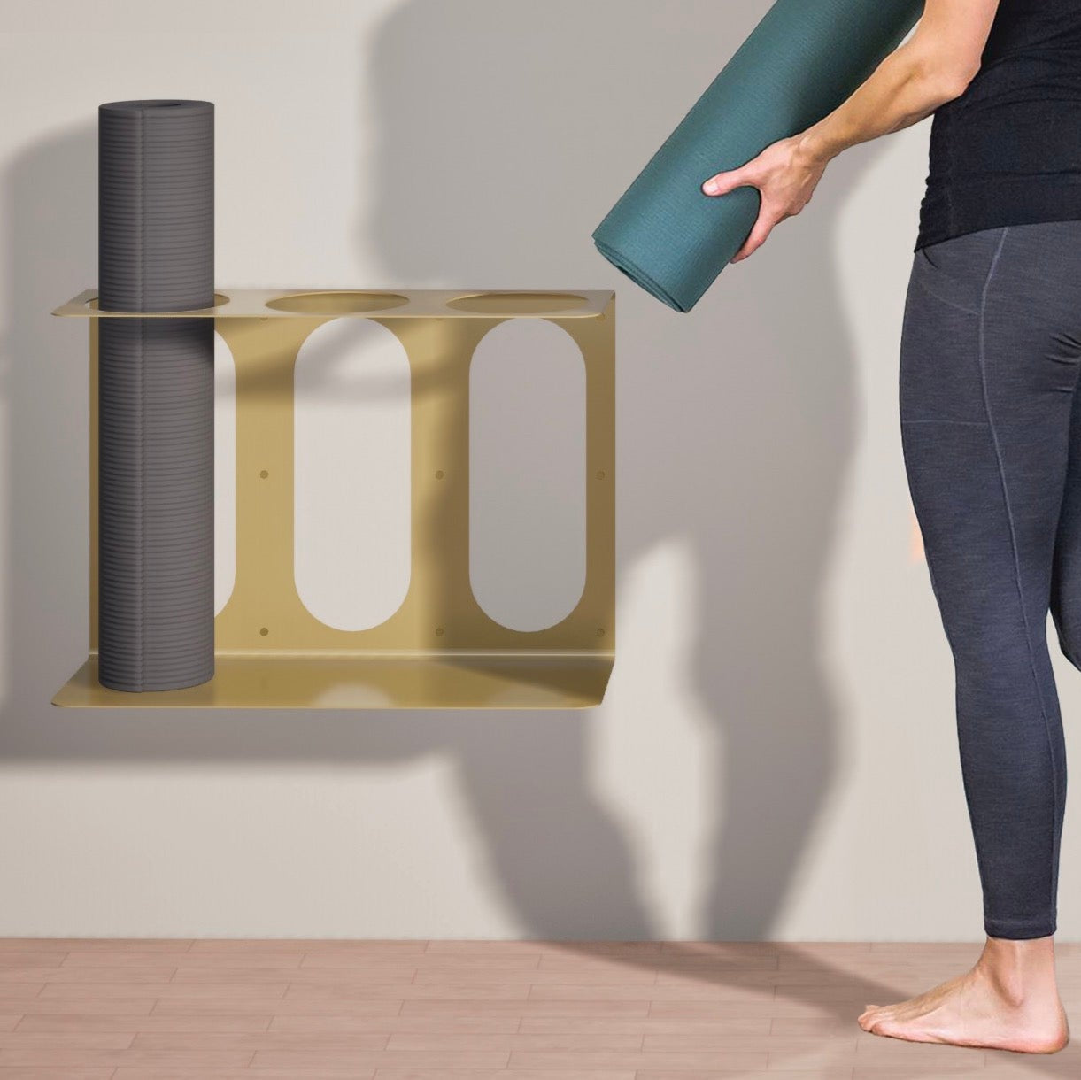 Person holding a yoga mat next to a gold yoga mat stand, attached to the wall with 3 places to hold mats.