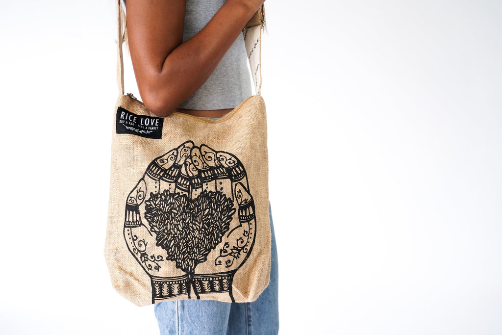 Beige tote bag with black grains in a heart pattern held by a persons hands on a white background