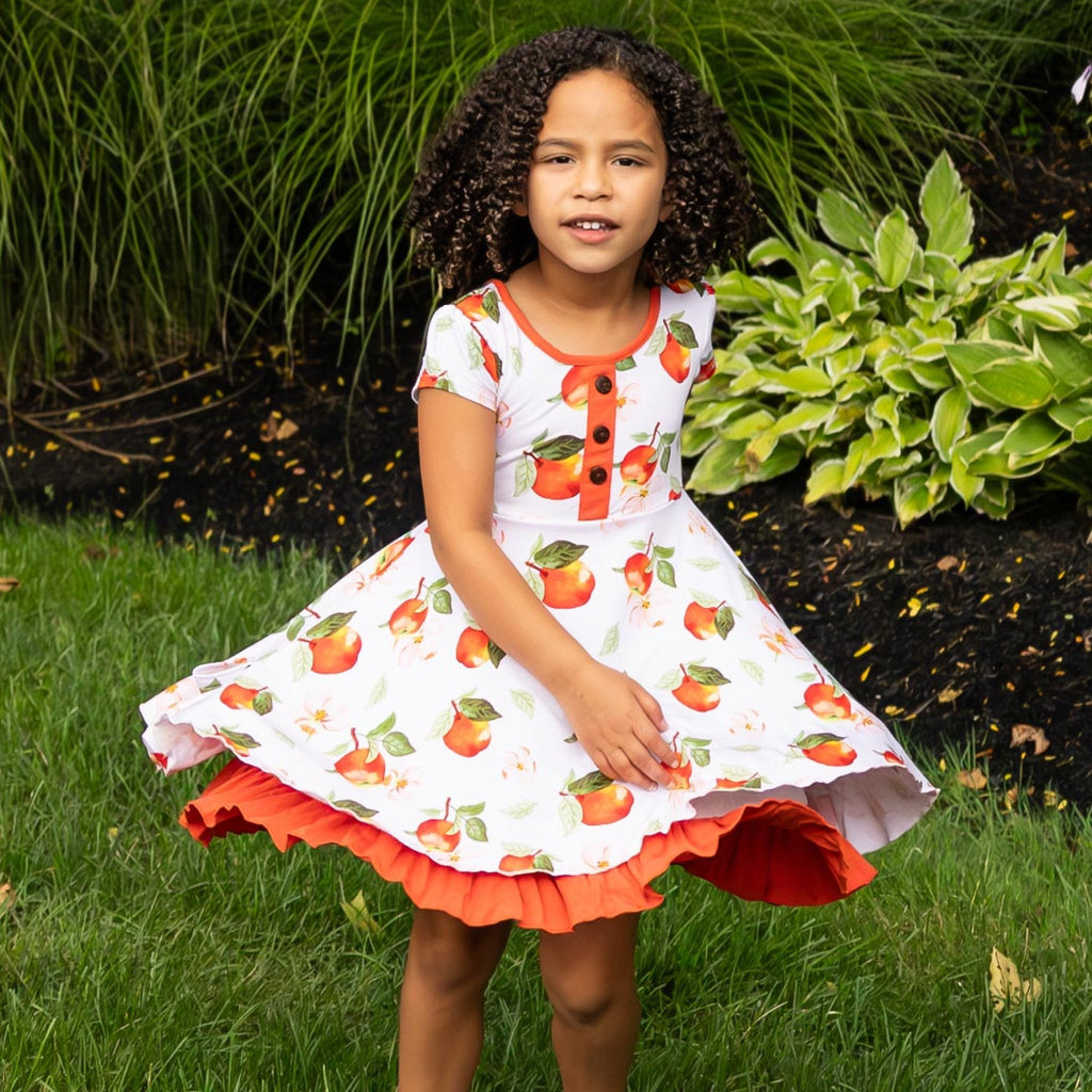 Young girl wearing a dress with apple pattern standing outdoors.