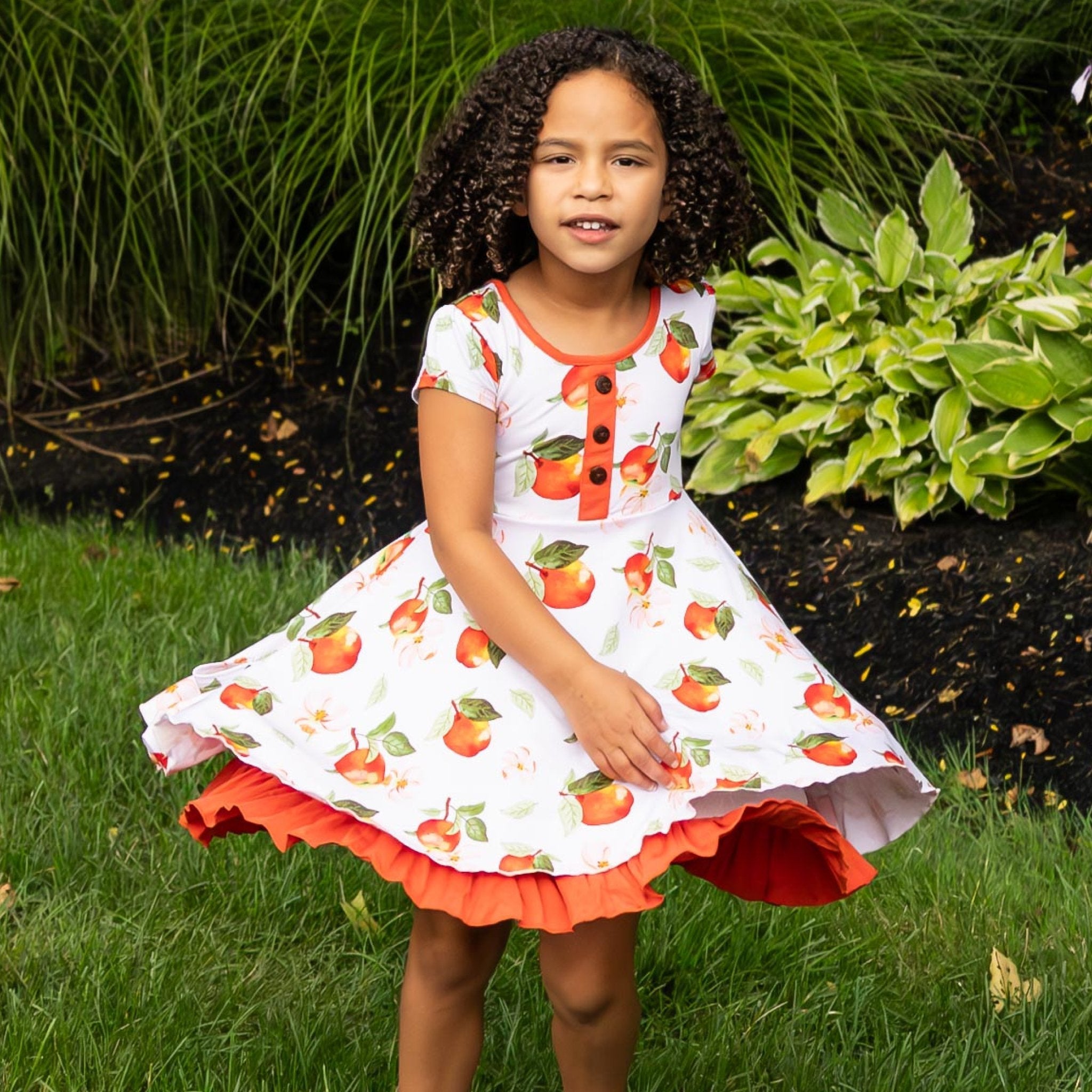 Young girl wearing a dress with apple pattern standing outdoors.