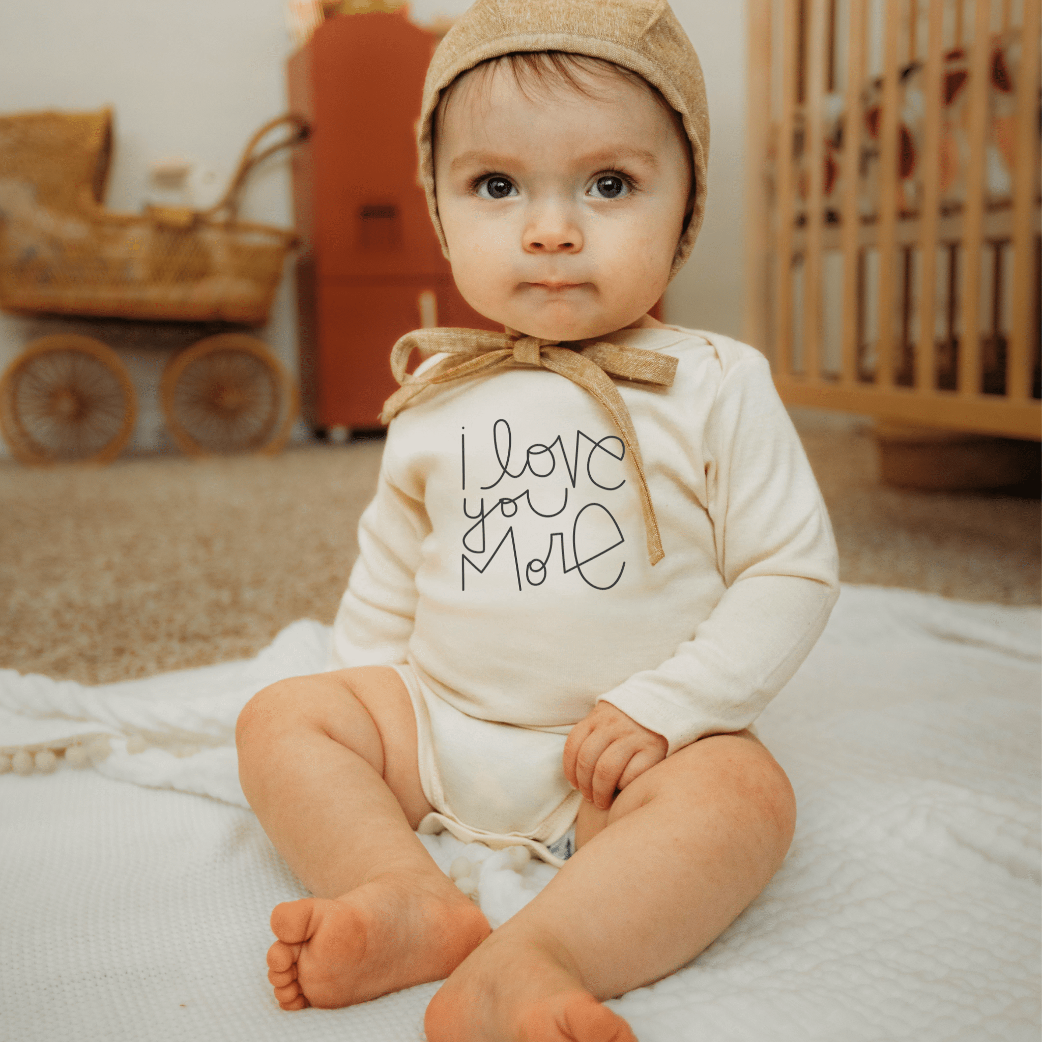 Baby wearing a 'Love You More' onesie and a brown bonnet in a nursery setting.