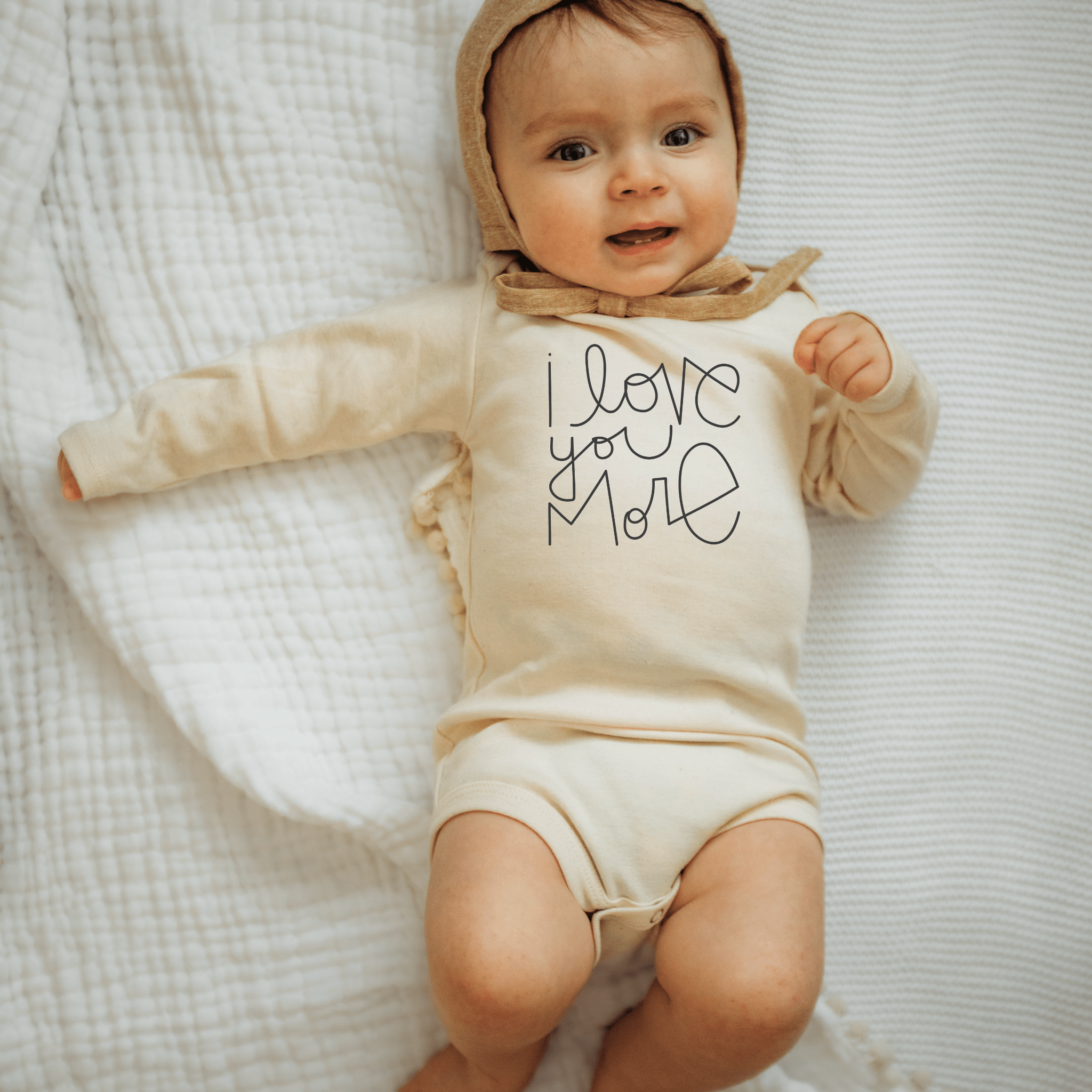 Baby wearing a beige onesie with 'I love you more' text on a white textured blanket.