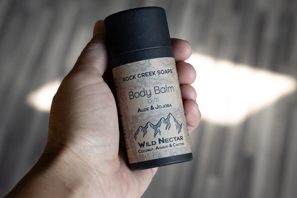 Hand holding a Rock Creek Soaps body balm 'Wild Nectar' container with a blurred background