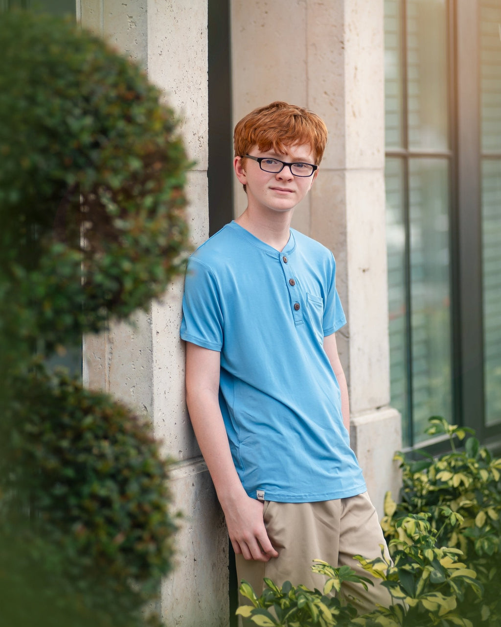 Person wearing a blue shirt leaning against a wall with greenery around