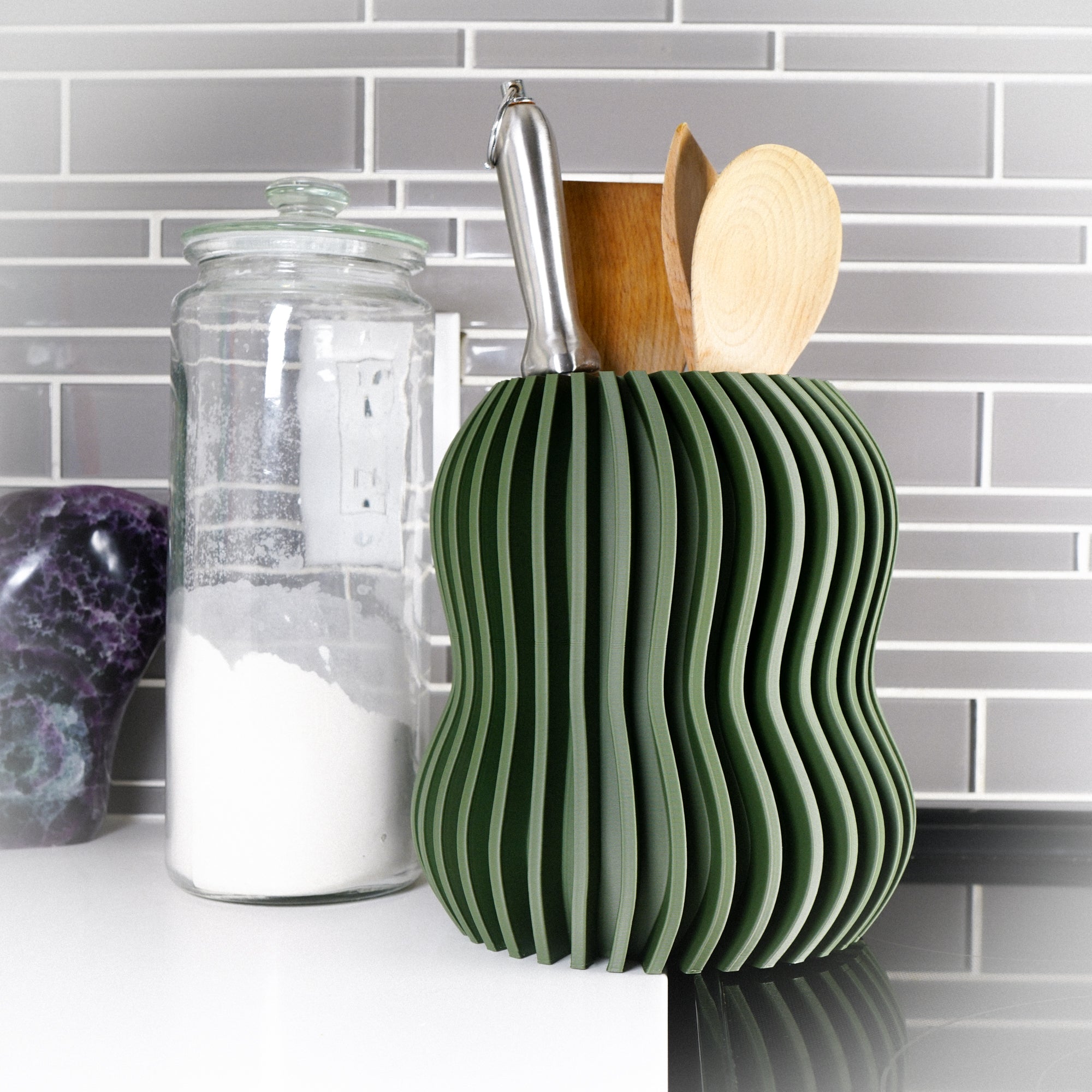 Green wavy kitchen utensil holder with wooden spoons and a metal ladle on a kitchen counter.