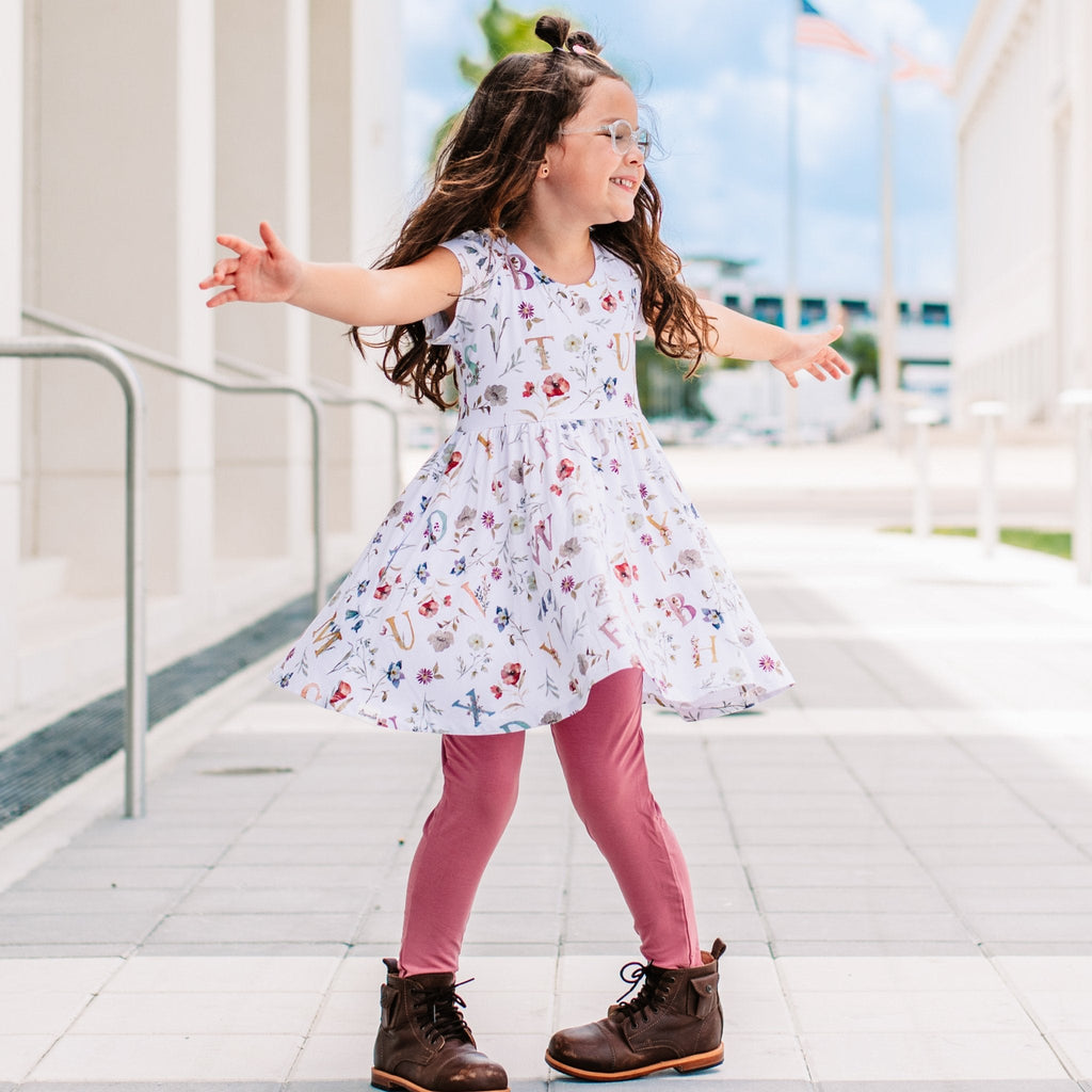 Young girl in a floral dress and pink leggings standing on a tiled floor with a blurred outdoor background.