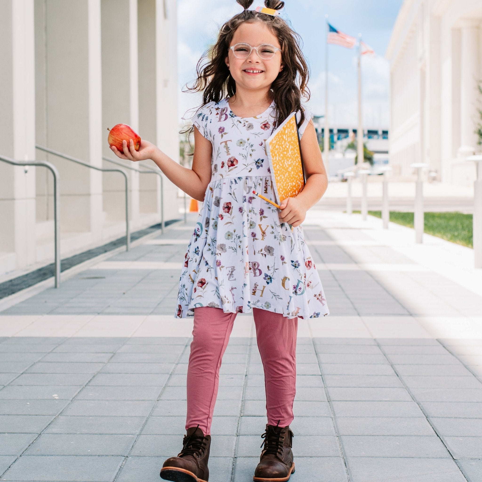 Young girl in a floral dress holding an apple and a yellow book outdoors.
