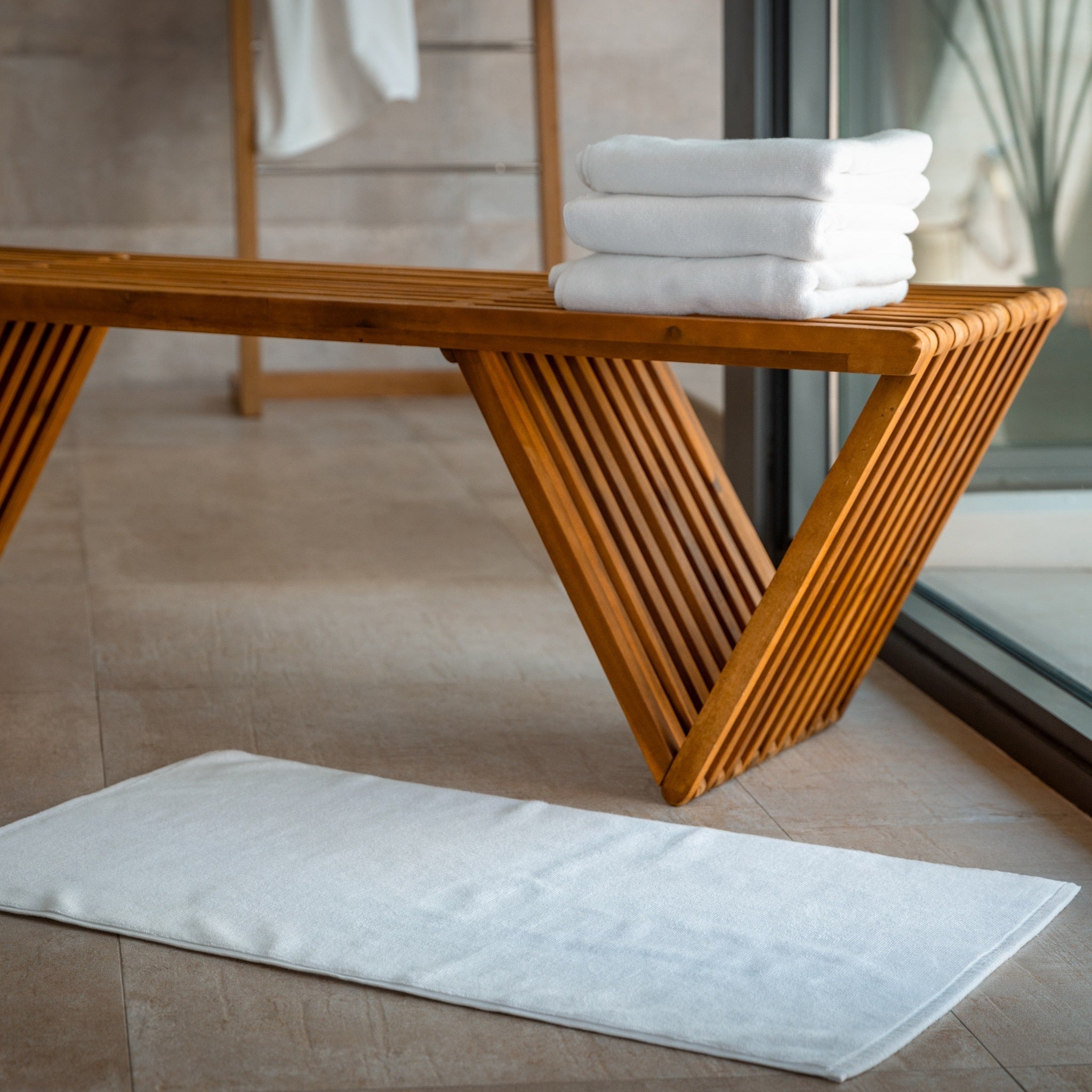 White bathmats stacked on a wooden bench with a white bath mat on the floor in front of it in a bathroom setting