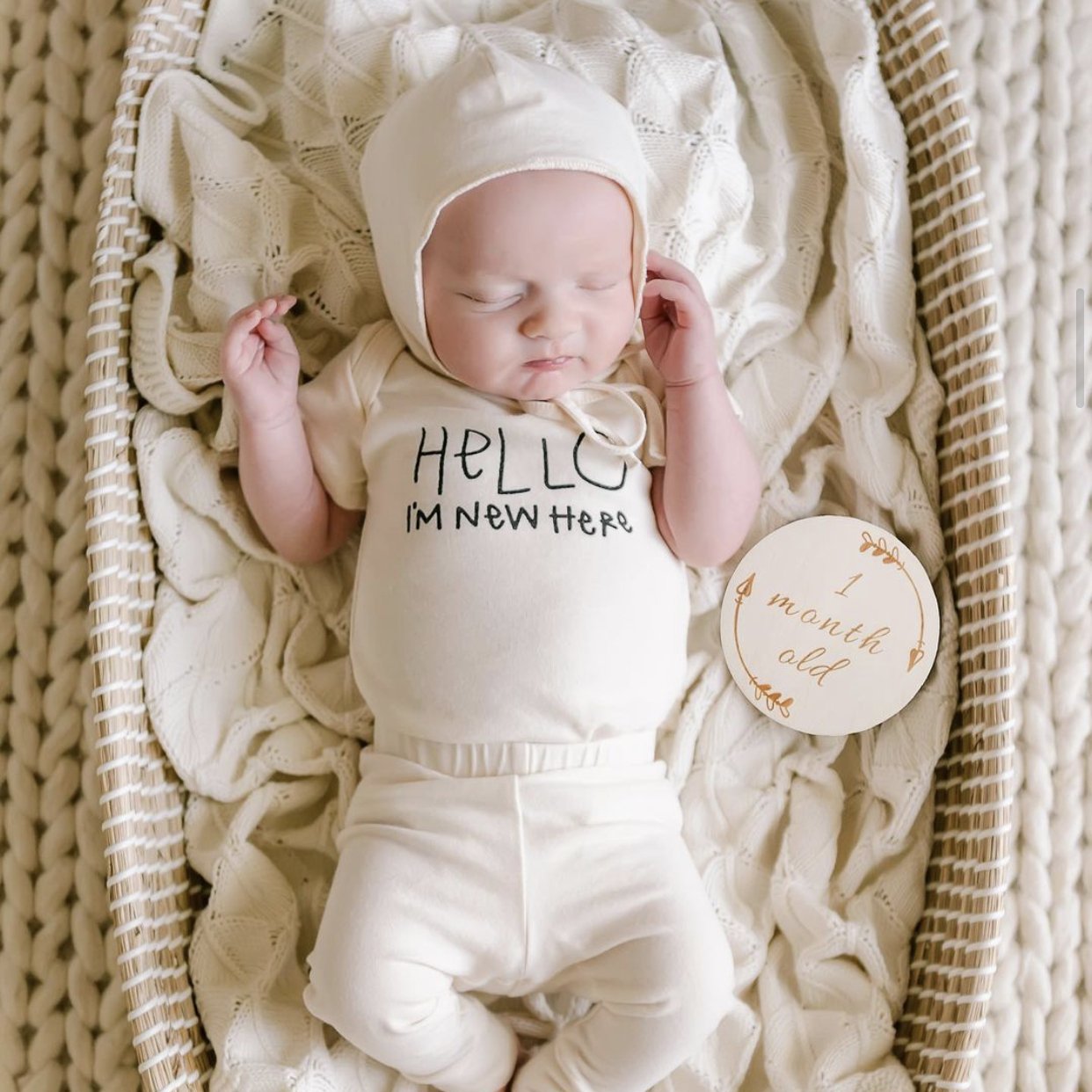 Newborn baby in a white outfit with 'Hello, I'm new here' text, lying on a textured blanket.