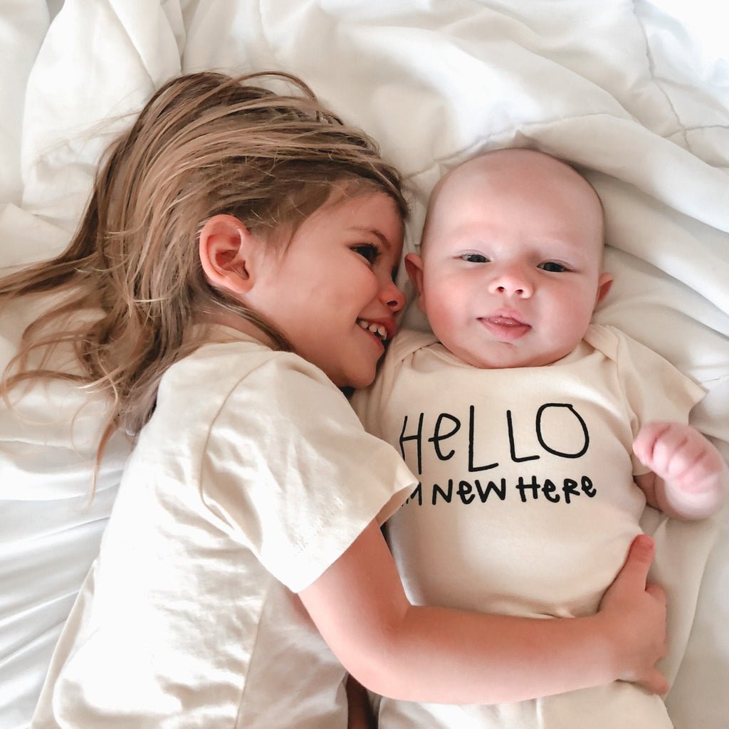 Two children lying on a white blanket, one wearing a 'Hello I'm new here' onesie.
