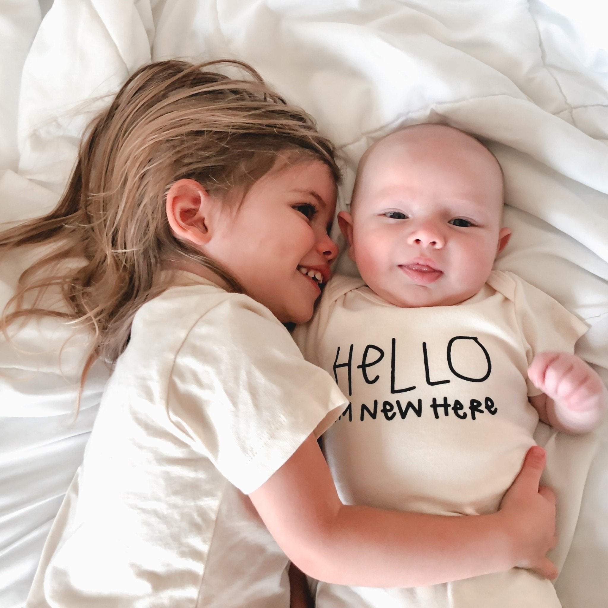 Two children lying on a white blanket, one wearing a 'Hello I'm new here' onesie.