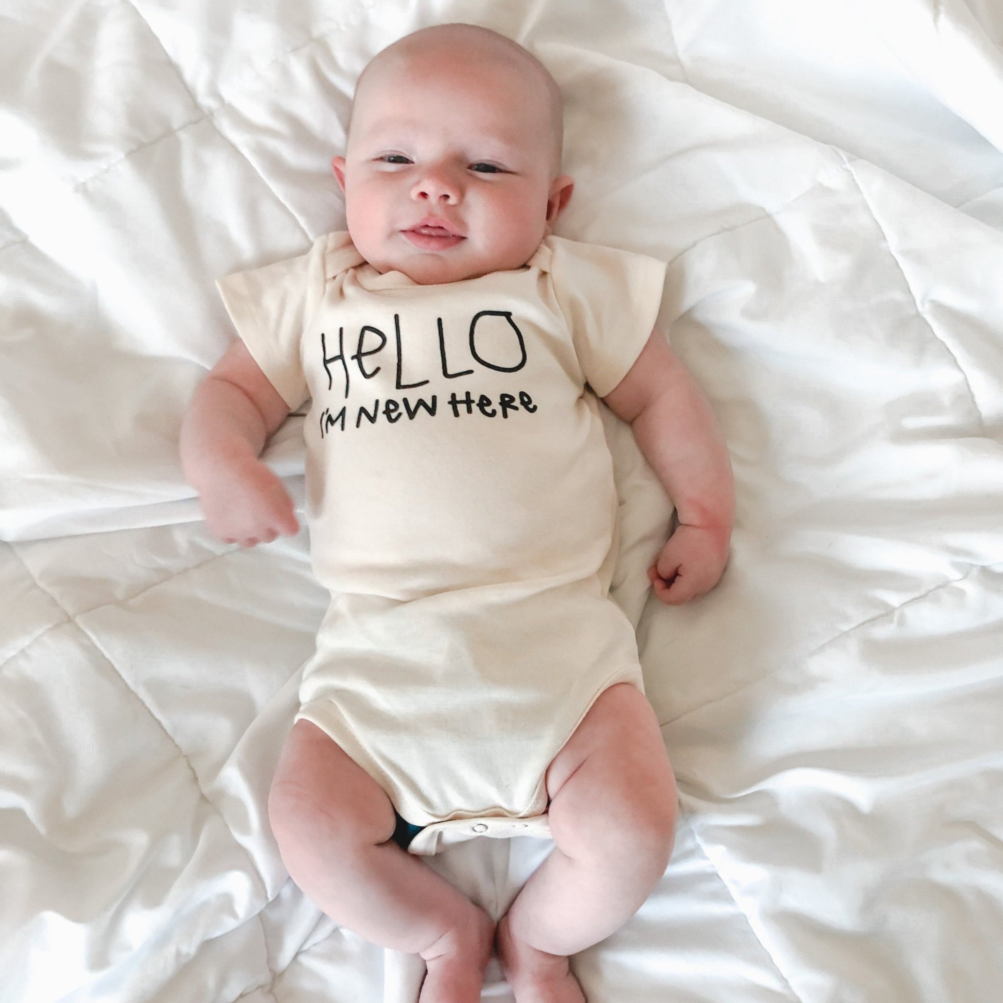 Baby wearing a onesie with 'Hello I'm new here' text, lying on a white blanket.