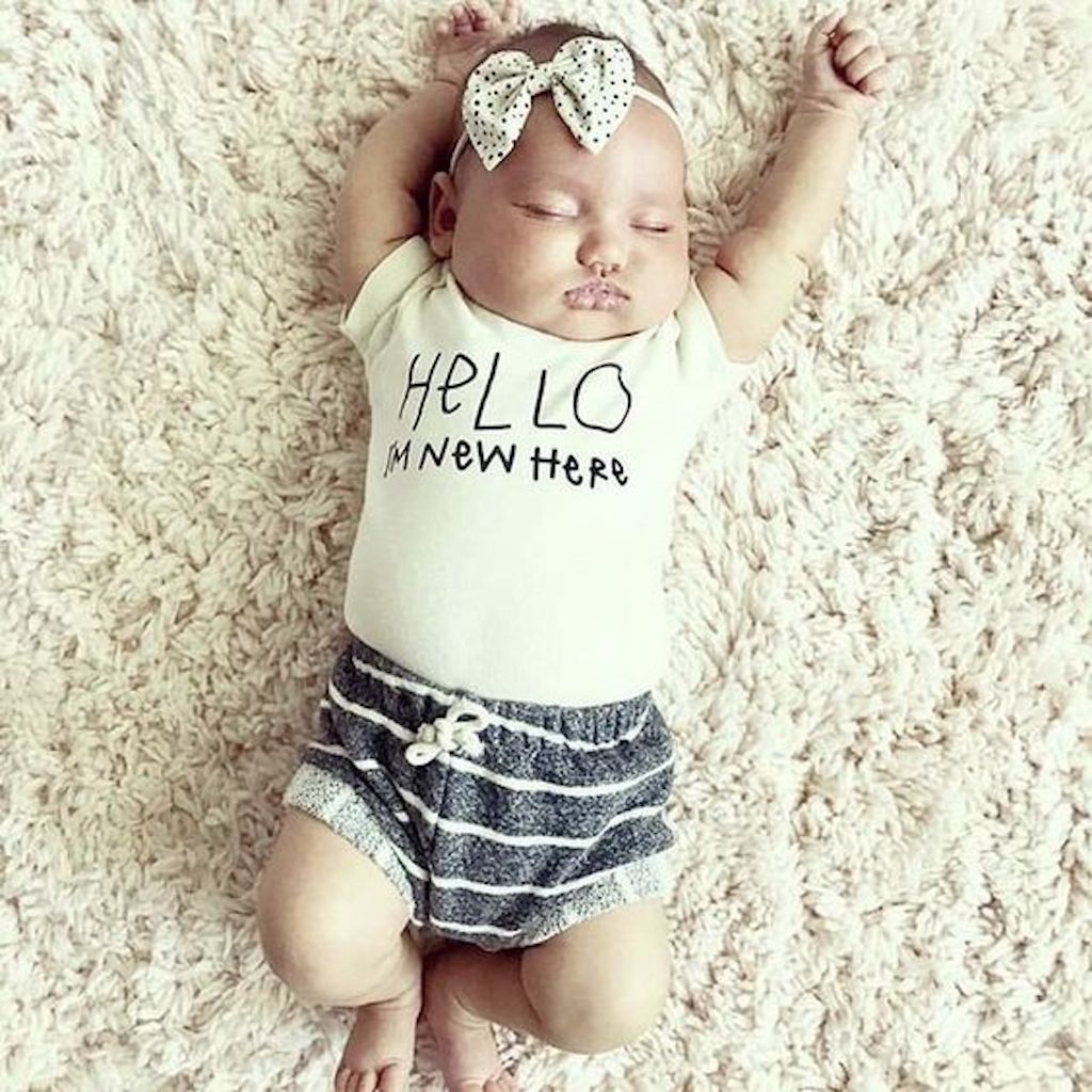 Newborn baby wearing a 'Hello I'm New Here' outfit on a soft, beige surface.