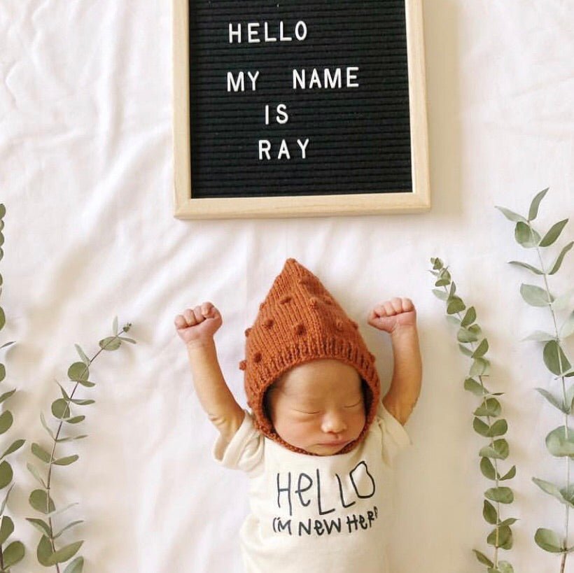 Newborn baby wearing a brown hat and white shirt with text, lying on a white blanket with green leaves.