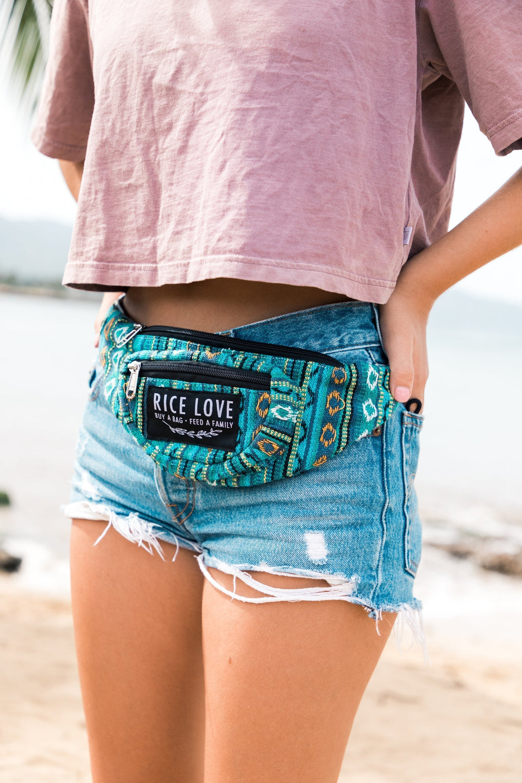 Person wearing a blue denim shorts and a patterned waist bag with 'Rice Love' branding on a beach.