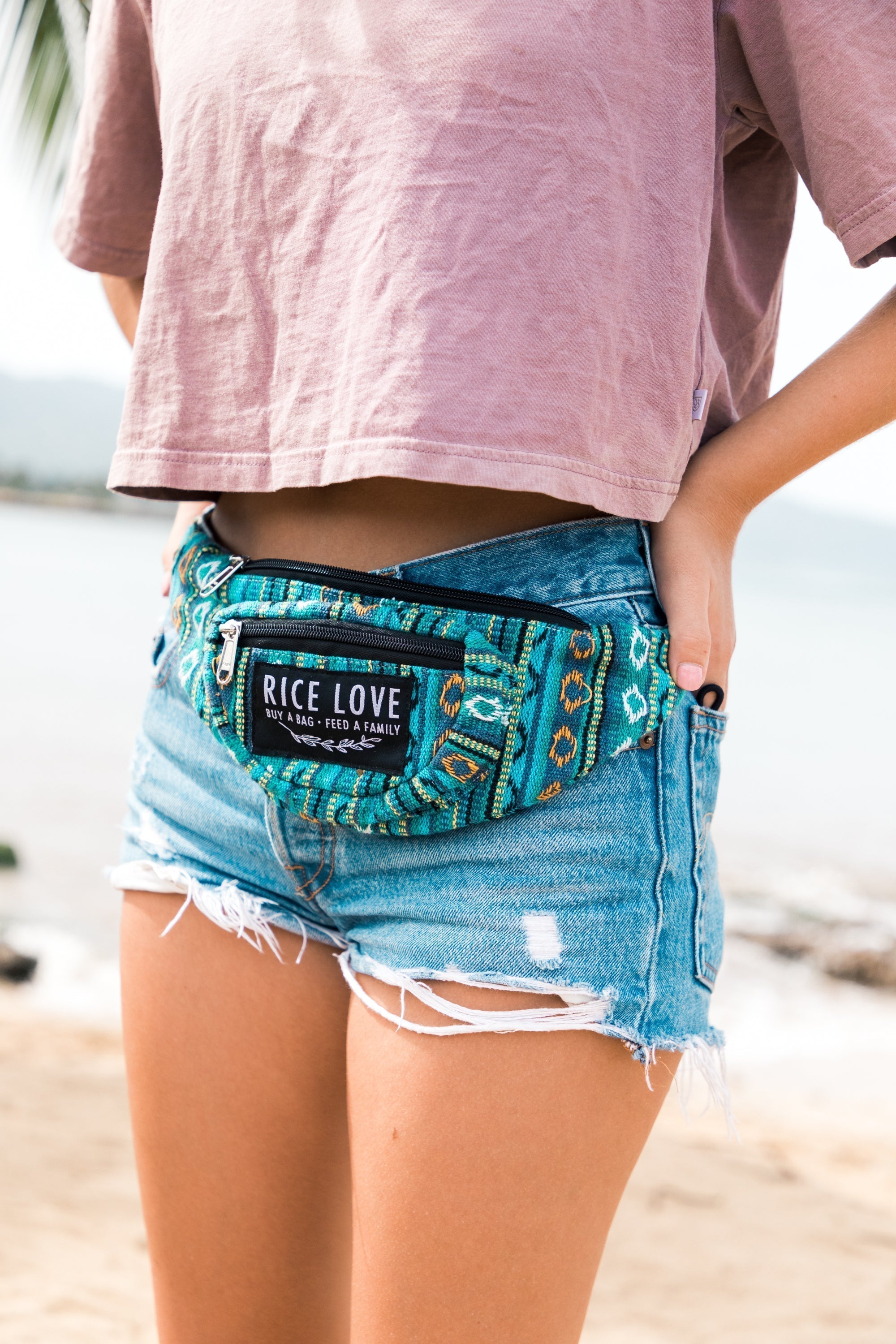 Person wearing a blue denim shorts and a patterned waist bag with 'Rice Love' branding on a beach.