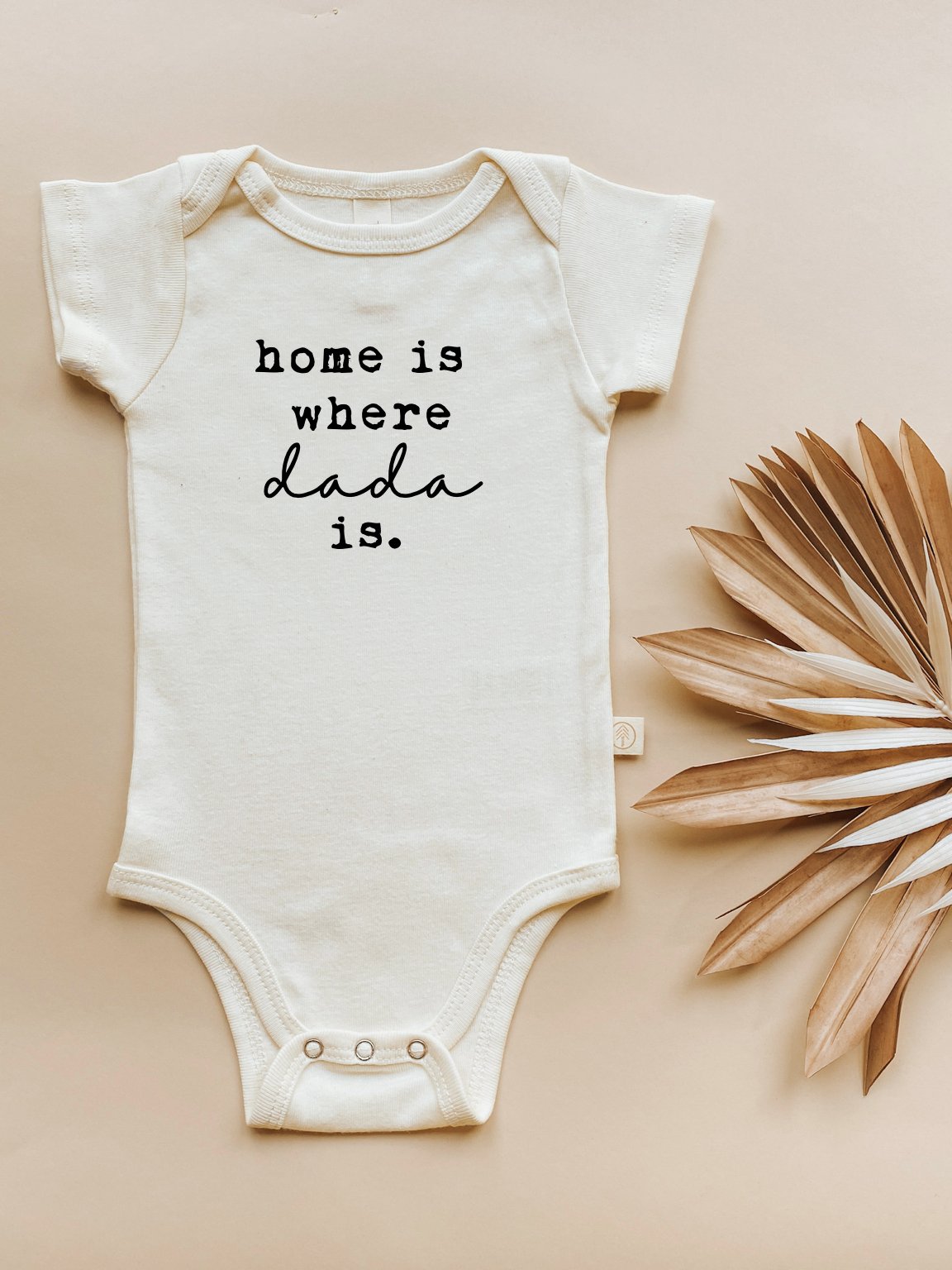 White baby onesie with 'home is where dad is' text on a beige background with a dried leaf.