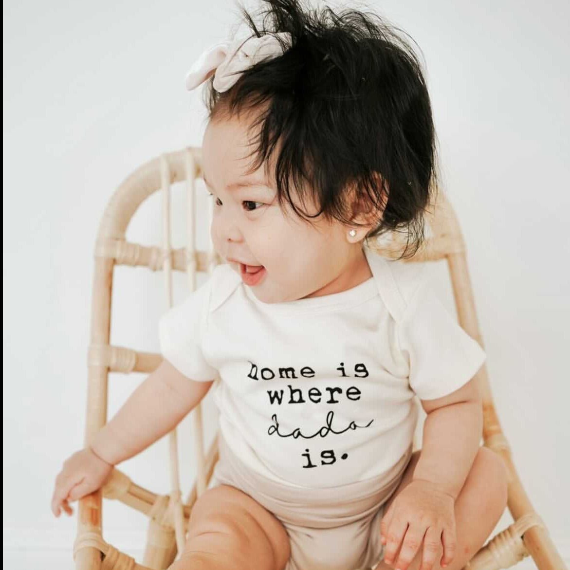 Baby wearing a onesie with 'Home is where dad is.' text, sitting on a wicker chair.
