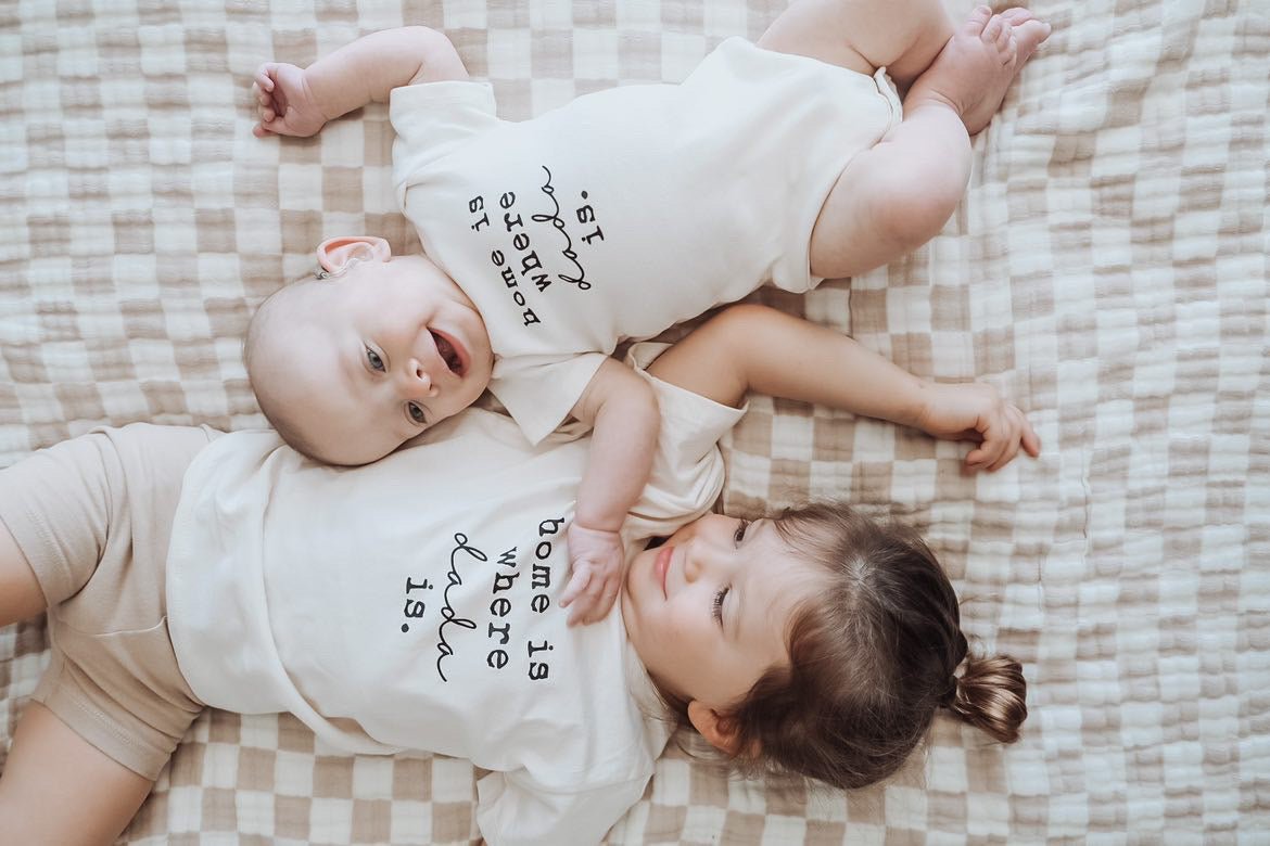 Two babies wearing matching outfits with text lying on a checkered blanket.