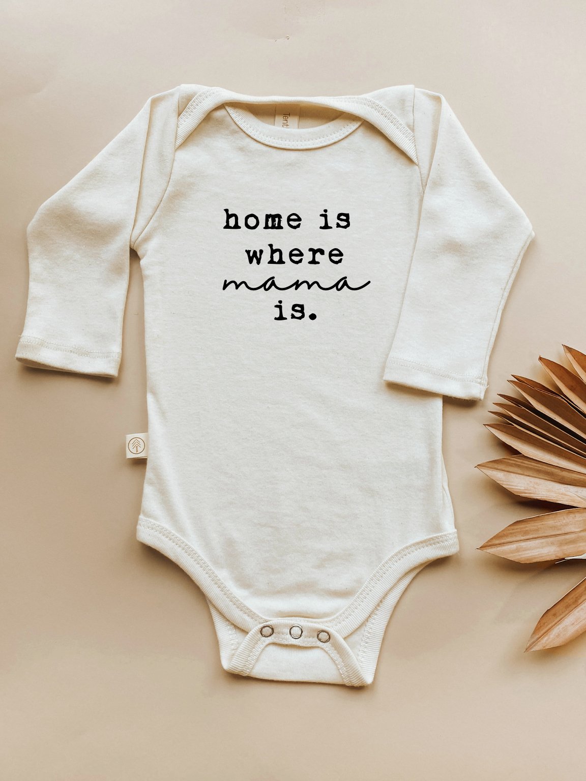 White baby onesie with 'home is where mama is' text on a beige background