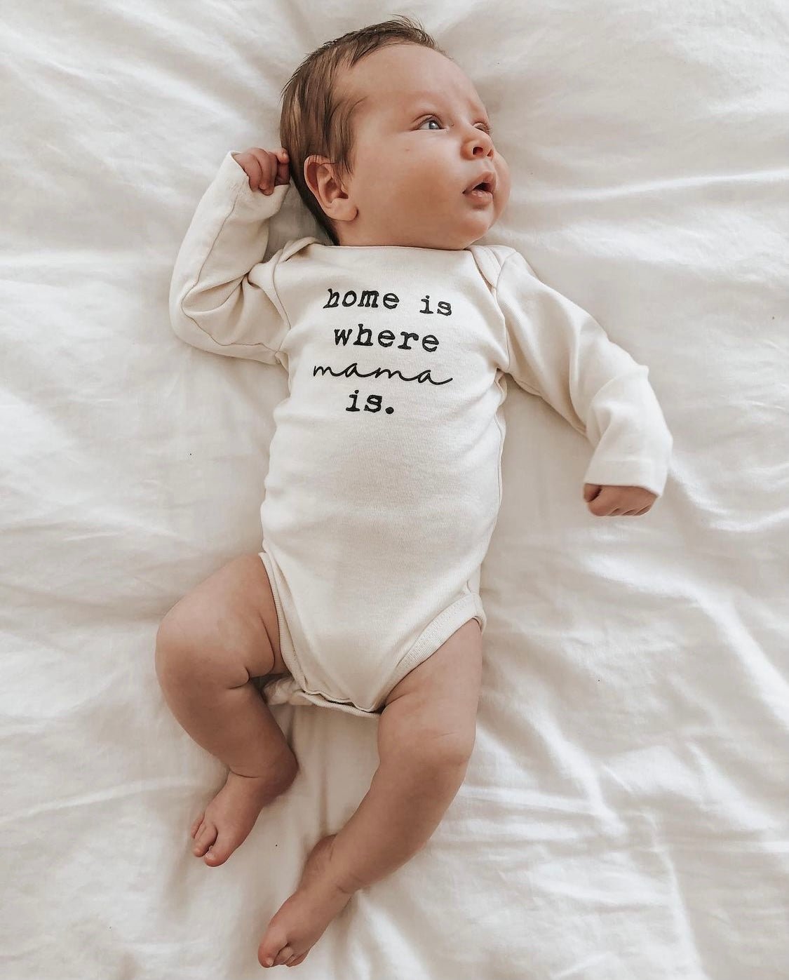 Baby wearing a onesie with 'home is where mama is' text, lying on a white surface.