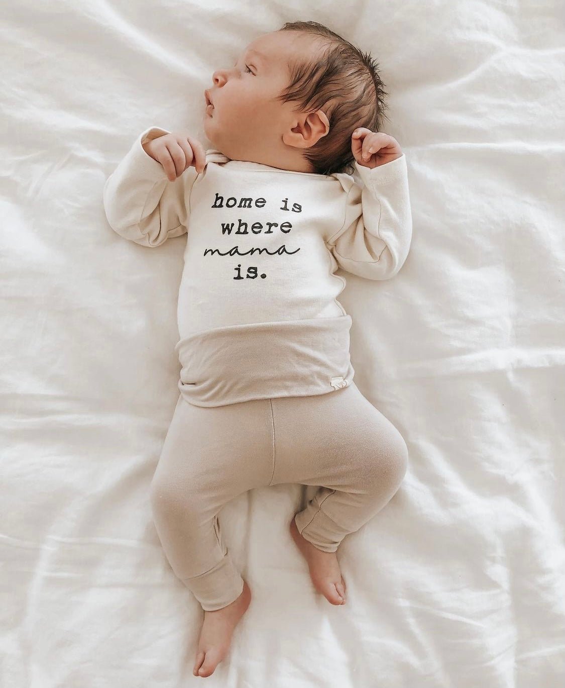 Newborn baby wearing a onesie with 'home is where mama is' text on a white blanket.