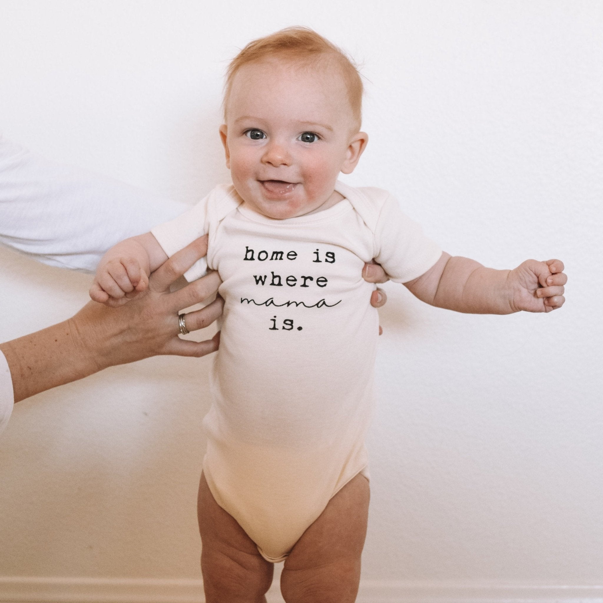 Baby wearing a onesie with 'home is where mama is' text, held by an adult against a white background