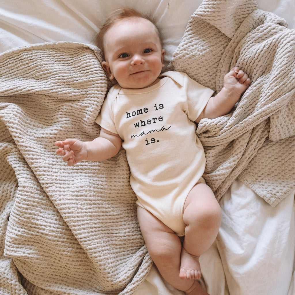 Baby wearing a onesie with 'home is where mama is' text, lying on a soft textured blanket.