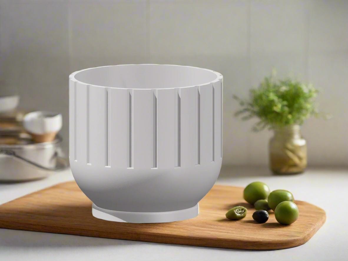 White plant pot on a wooden cutting board with olives and a vase in the background