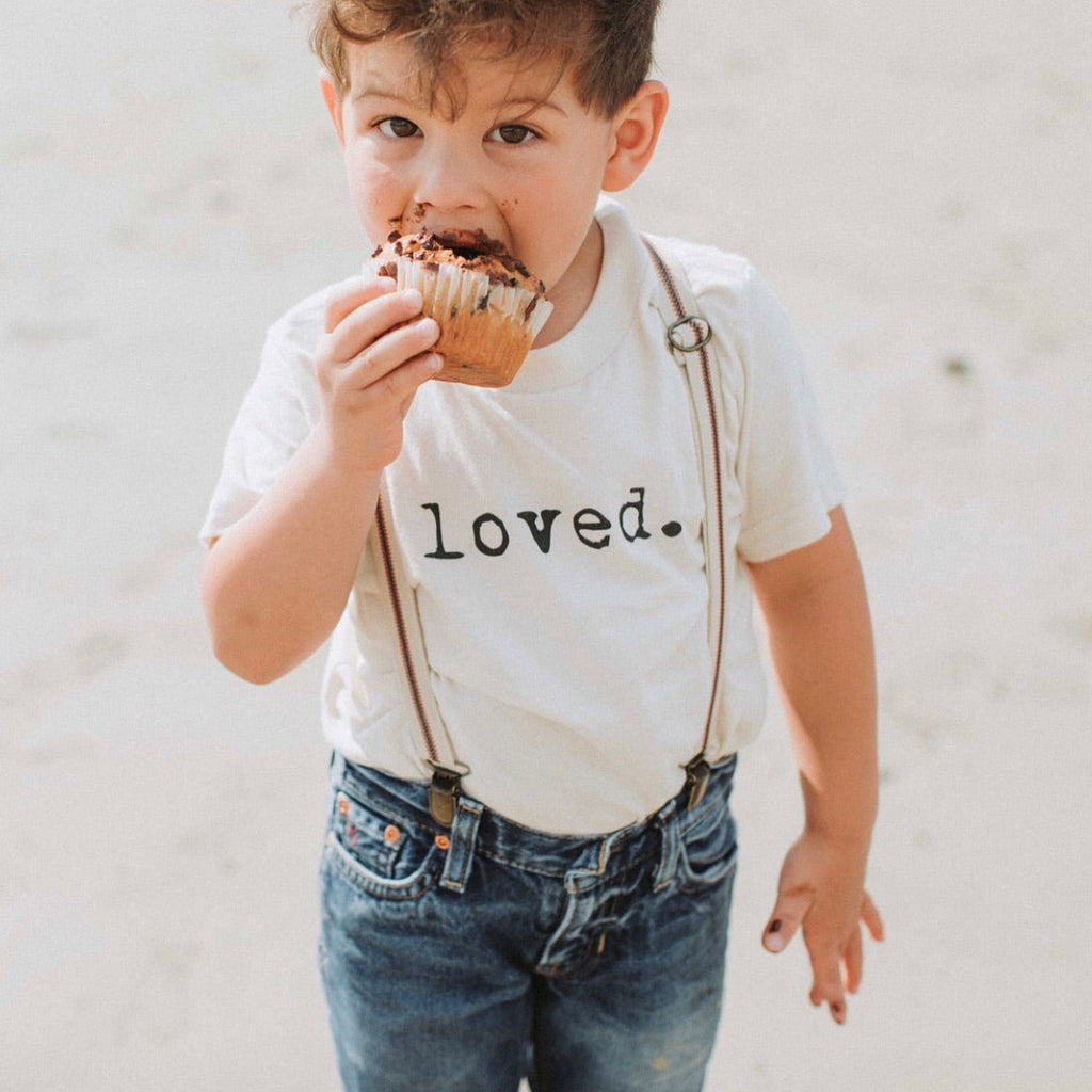 Child wearing a shirt with 'loved.' eating a muffin outdoors.